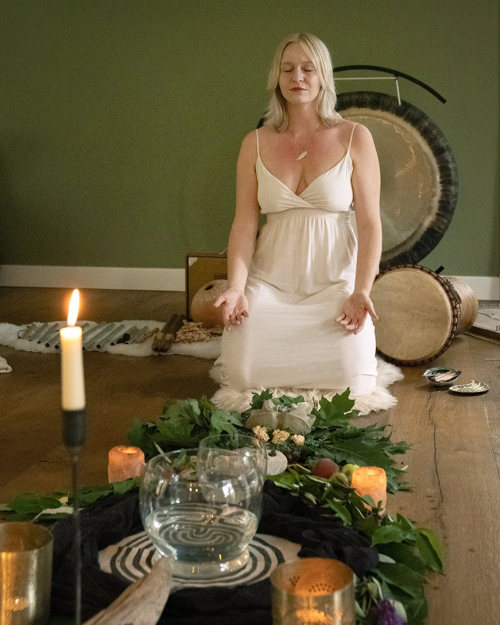 A woman with blonde hair and closed eyes sits cross-legged on a fur rug with her hands resting on her knees, surrounded by candles and greenery on a wooden floor, with musical drums and a gong in the background.