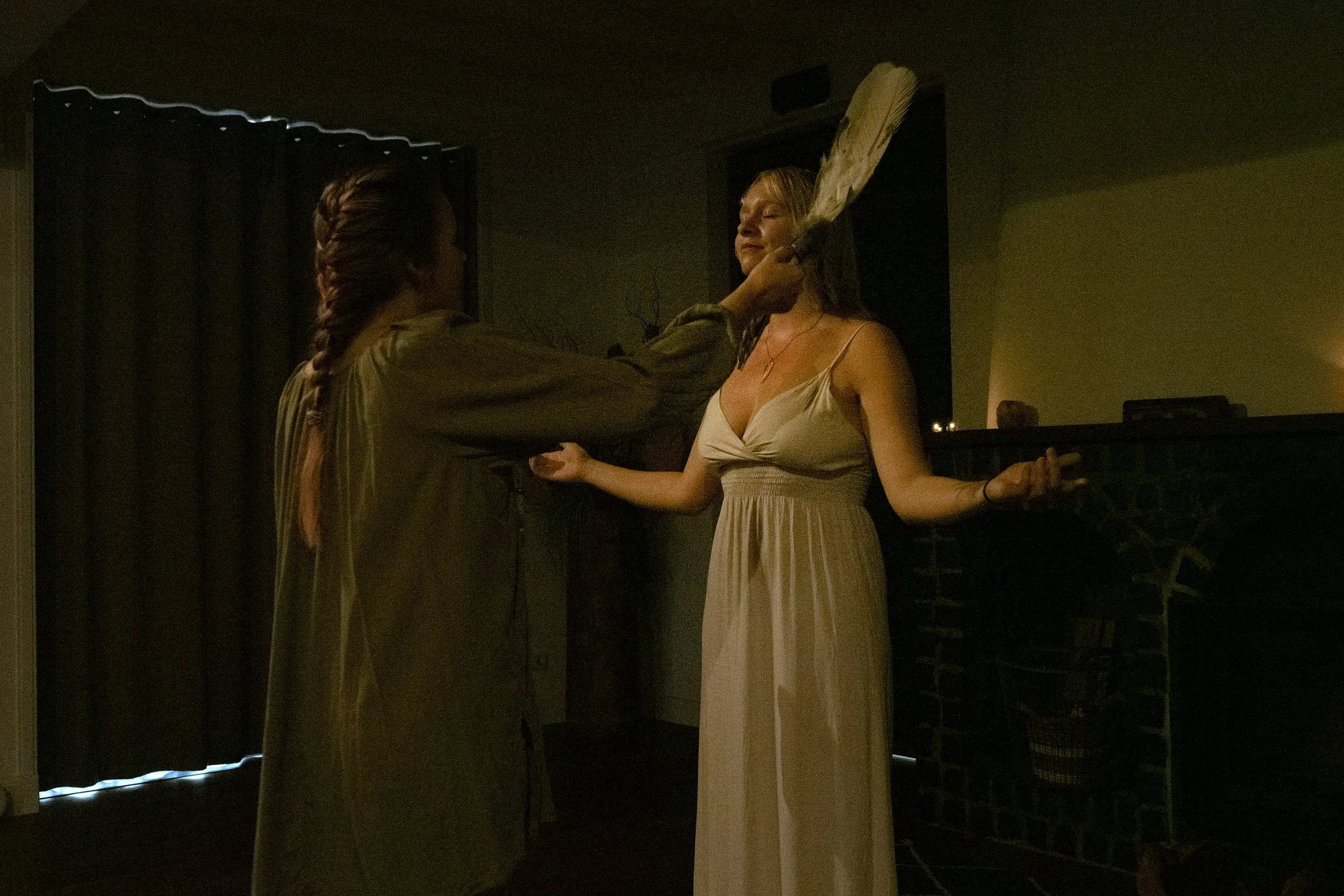 A woman in a light-colored dress with her eyes closed, being doused with a bucket of water by another woman in a green dress, in a dimly lit room.