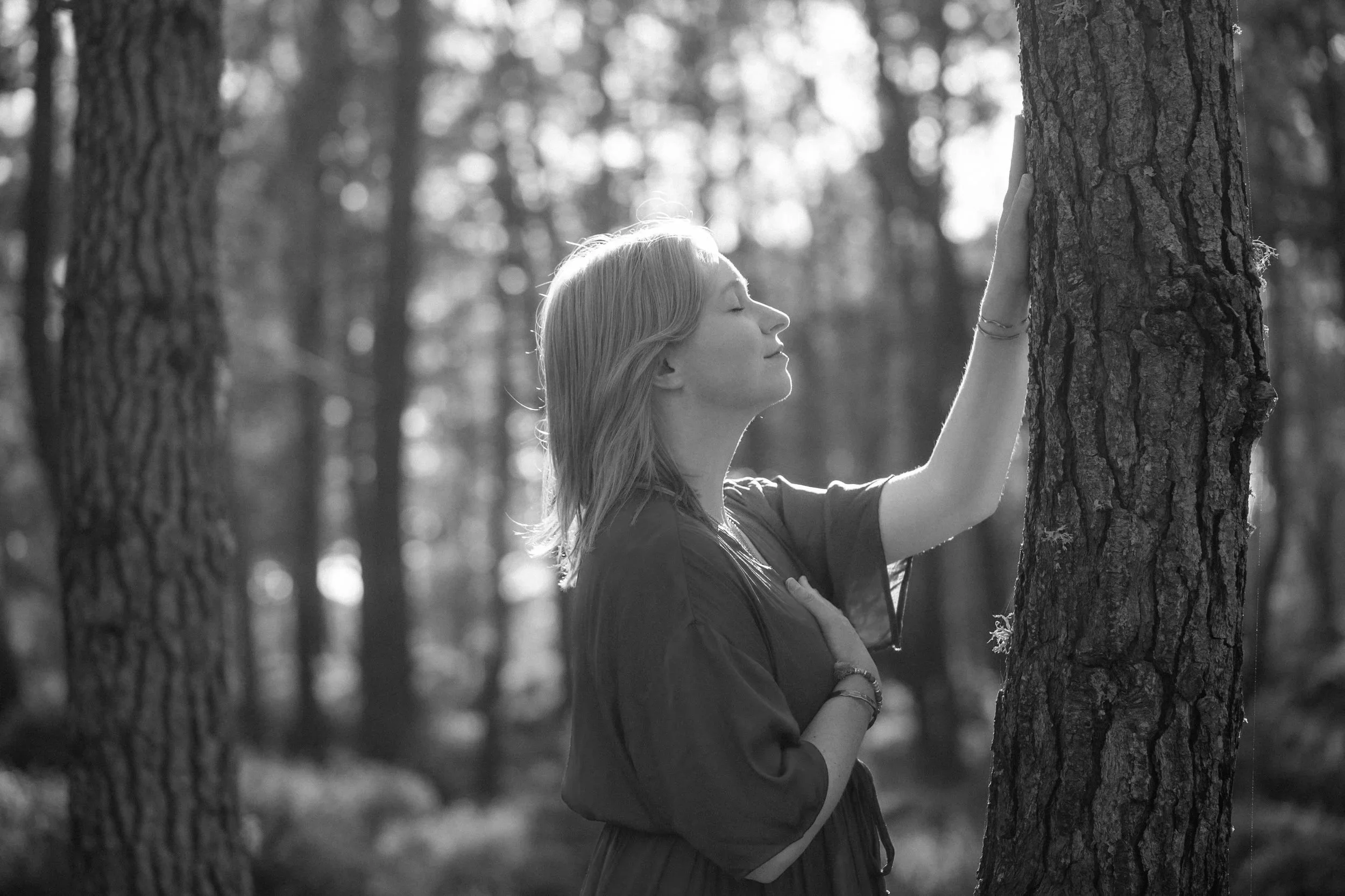 A woman with long hair and a bracelet touches a tree in a forest, eyes closed, appearing peaceful, in black and white.
