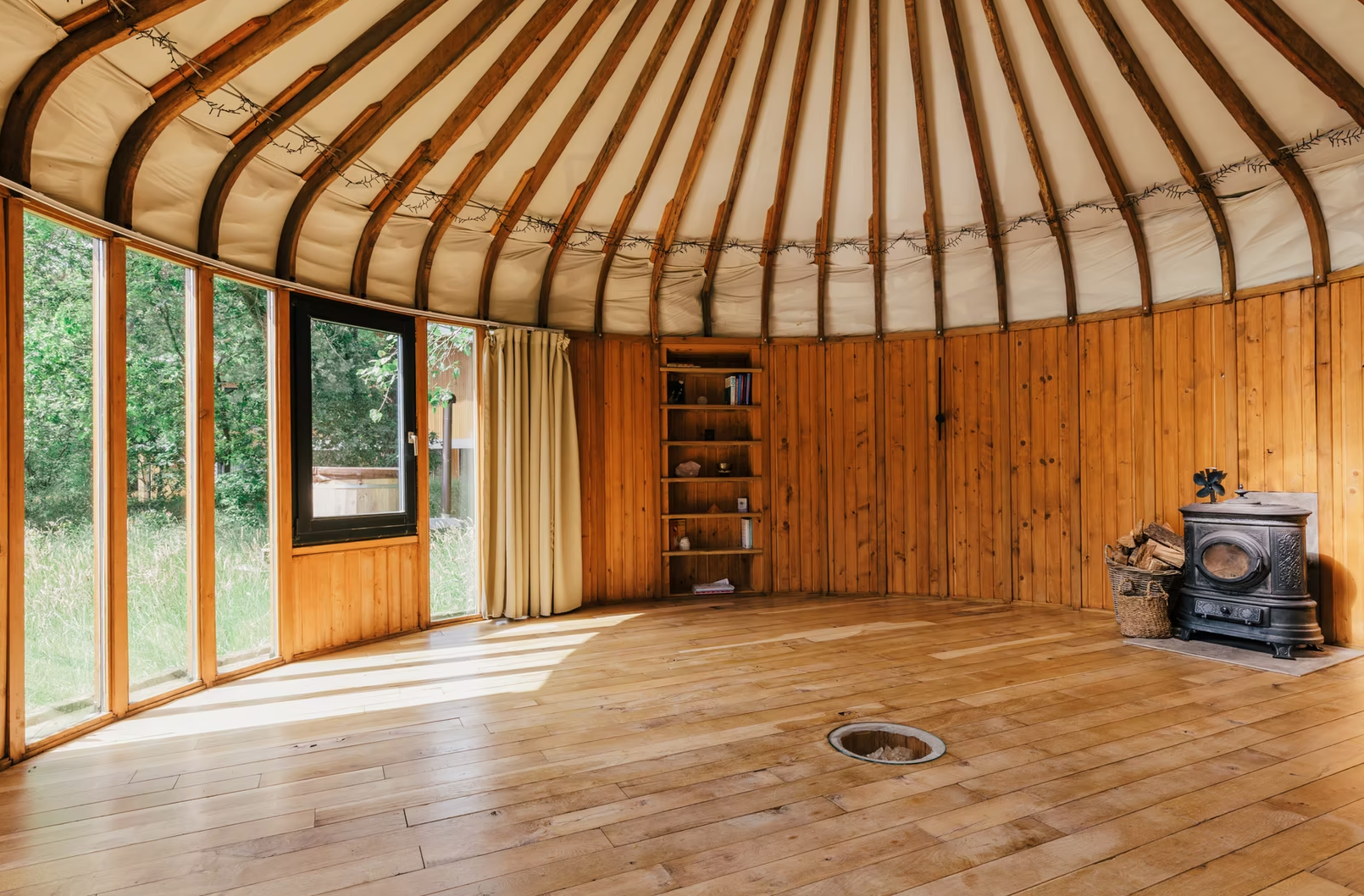 A circular wooden yurt interior with a curved ceiling, large windows along one wall, and a wood stove in the corner with a basket of firewood beside it.