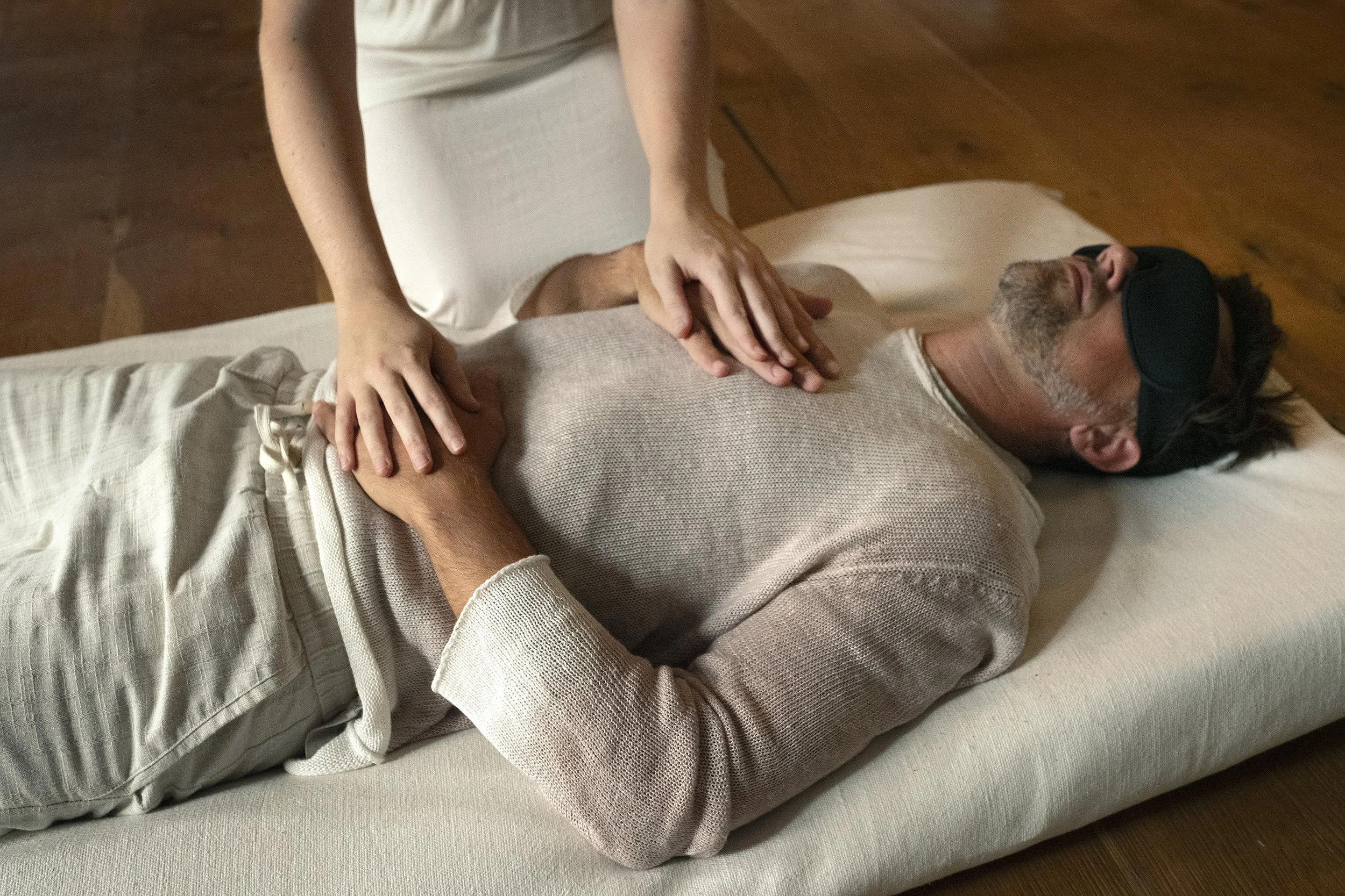 A person lying on a bed with white sheets, wearing a beige sweater, receiving a massage from another person whose hands are on their chest. The person on the bed is wearing black sleep goggles and has gray hair and a beard.