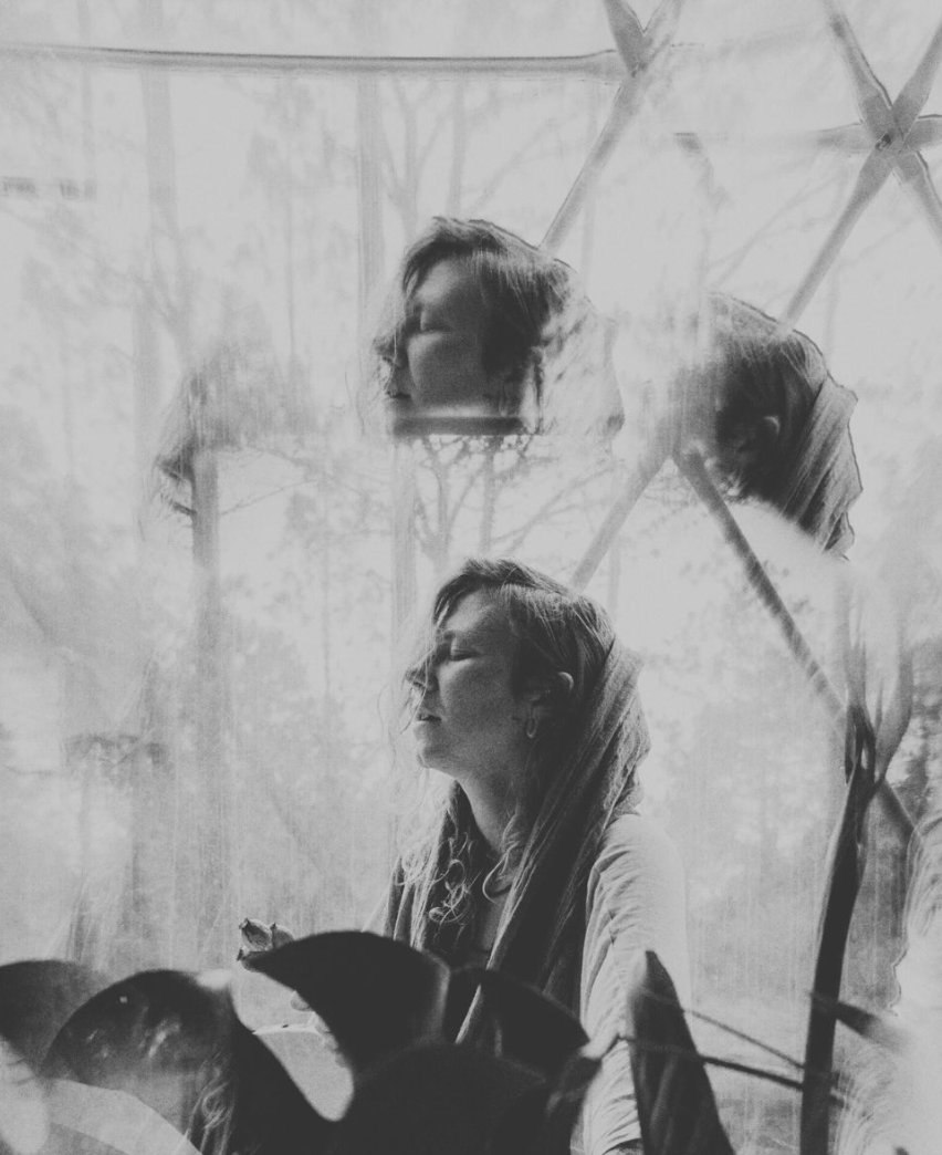 A black-and-white photo of a woman with wavy hair looking to the side, reflected multiple times in a glass or mirror, creating a layered effect.