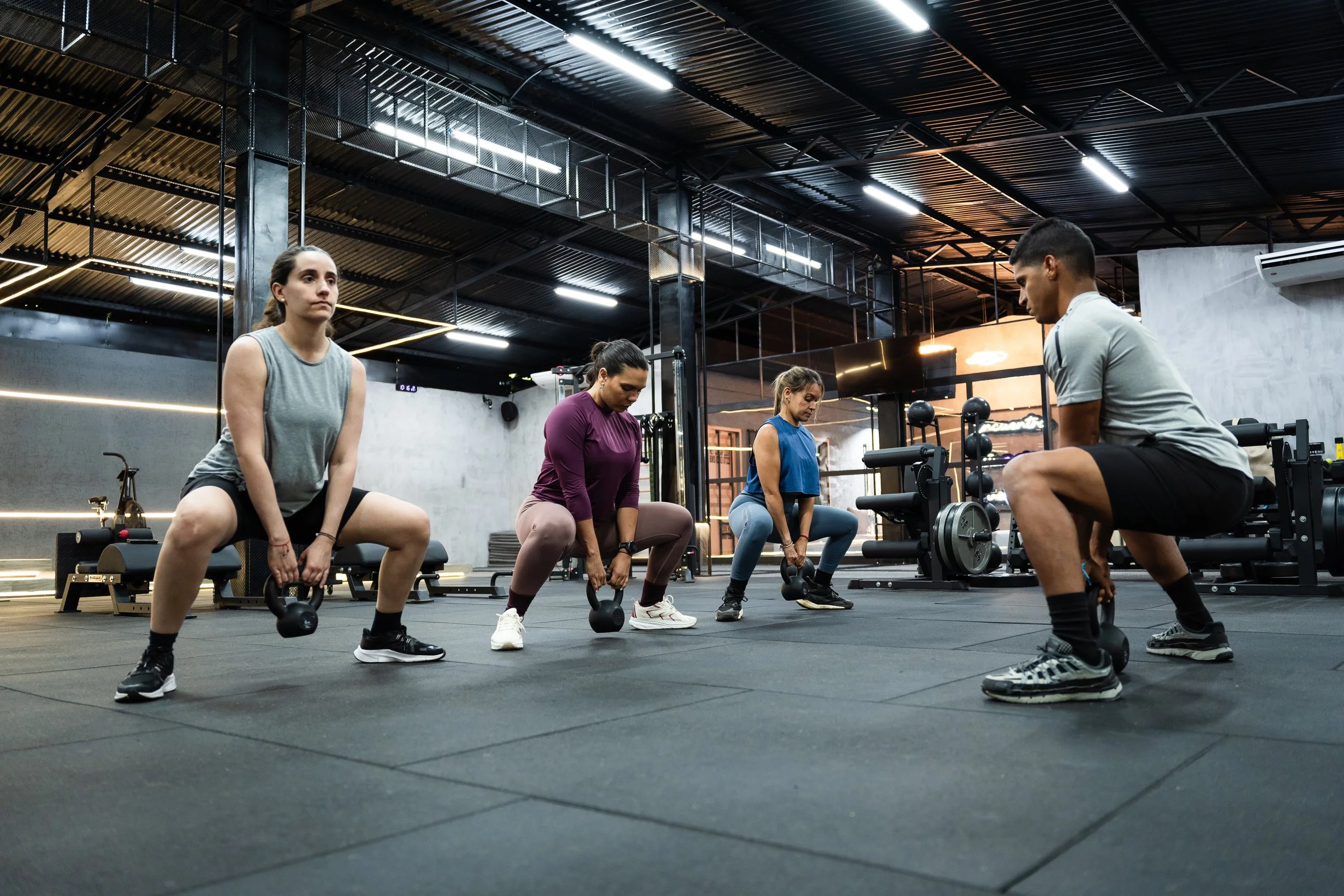 Four people in athletic wear performing squats with kettlebells in a gym.