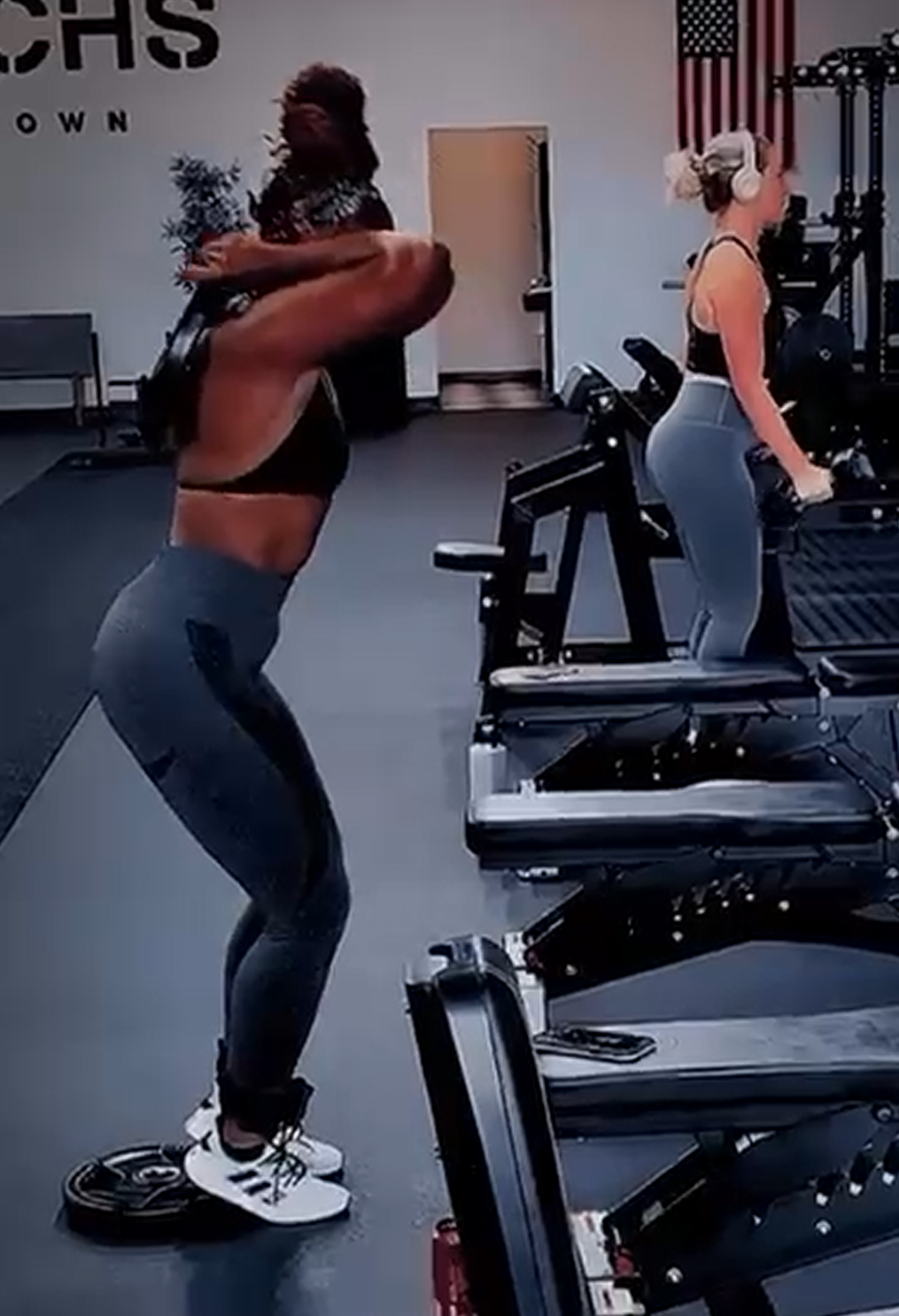 A woman lifting dumbbells during a workout at a gym, with another woman in the background using gym equipment, American flag decor on the wall.