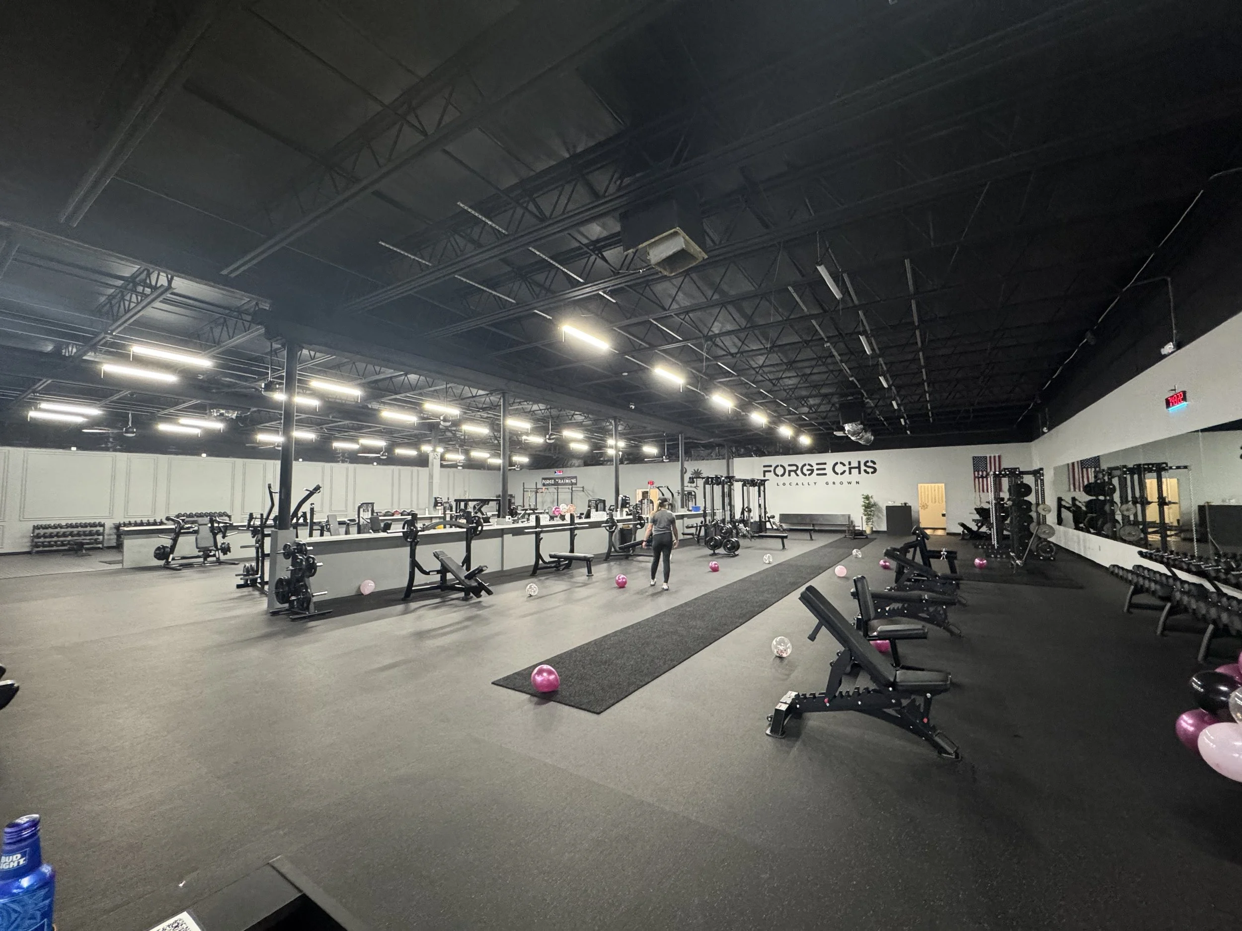 Empty gym with workout equipment, pink and clear exercise balls, black benches, weight racks, and a large wall sign that says 'FORGE CHS'. Bright overhead lights and a speaker hanging from the ceiling.