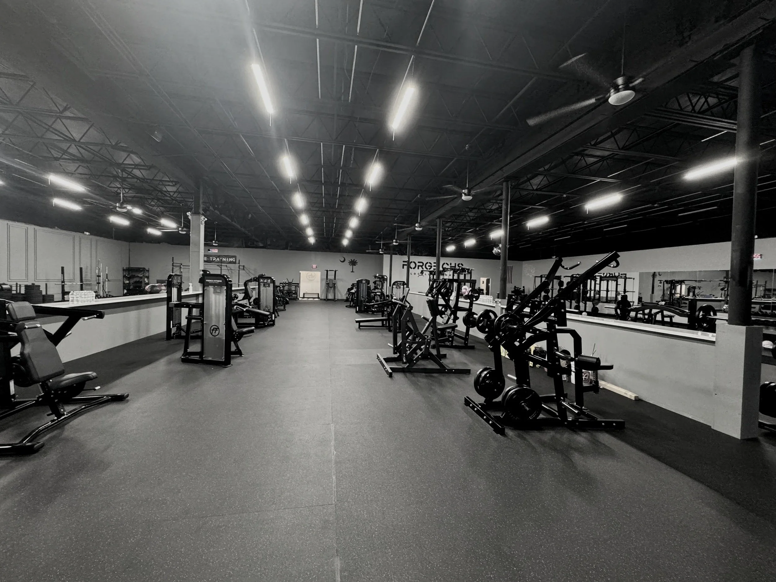 Empty gym with various workout equipment including weight machines, benches, and free weights, with a black ceiling and bright overhead lighting.