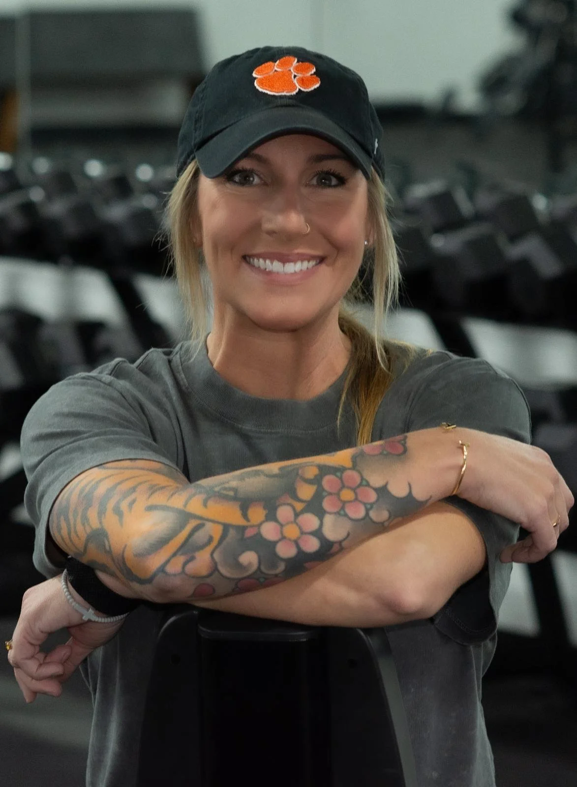 A smiling woman with long blonde hair, a nose piercing, and tattoos on her right arm, wearing a black cap with an orange paw print, a grey t-shirt, and a gold ring on her finger, sitting in a gym with black equipment in the background.