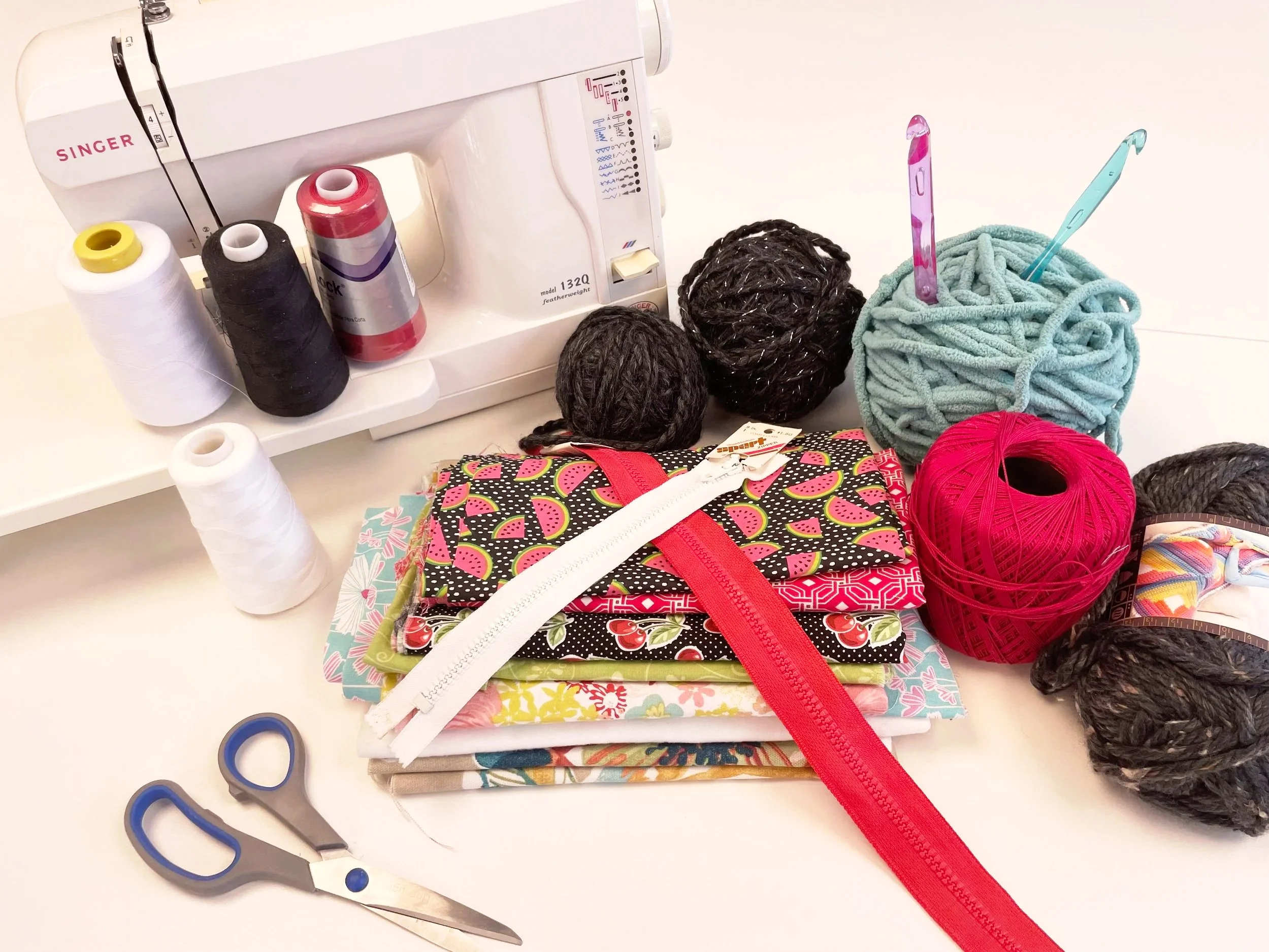 an array of fiber arts materials and supplies like a stack of multicolored fabric, different skeins of yarn and pools of thread, scissors, sewing machine, zippers