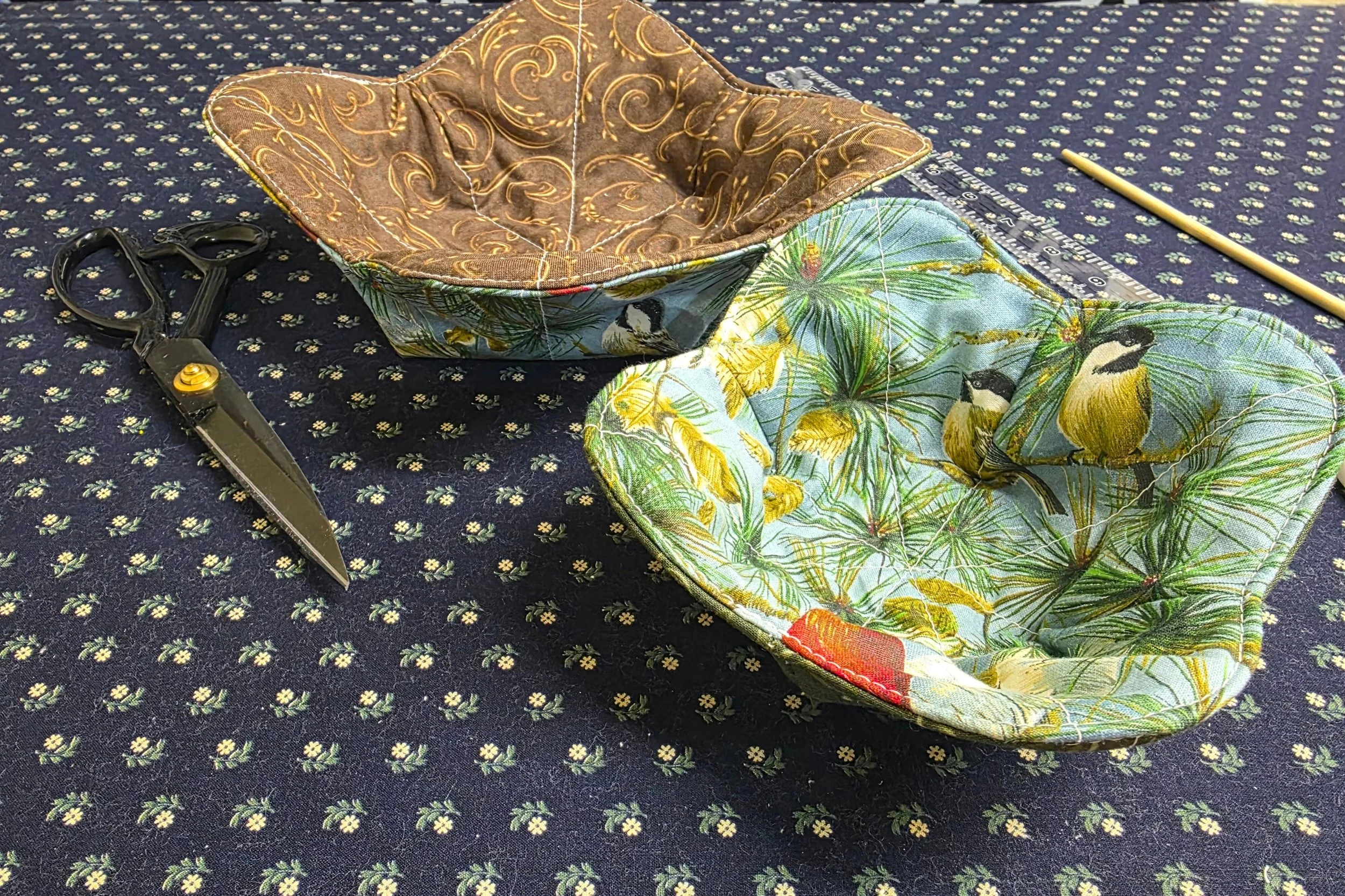 two bowl cozies with outdoorsy patterns atop a blue fabric pattern next to fabric scissors and a ruler