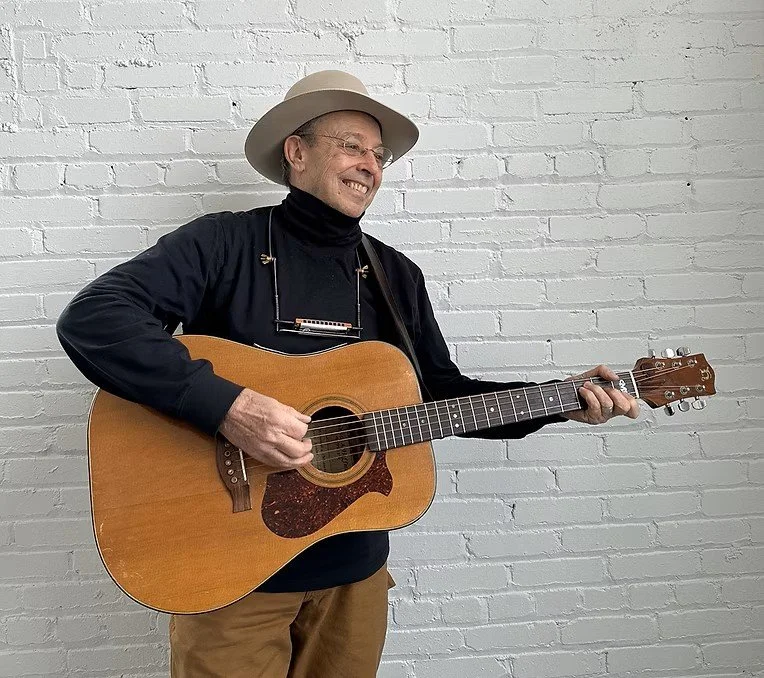 a photo of musician Charlie Maguire playing guitar and smiling while wearing a hat and a harmonica around his neck