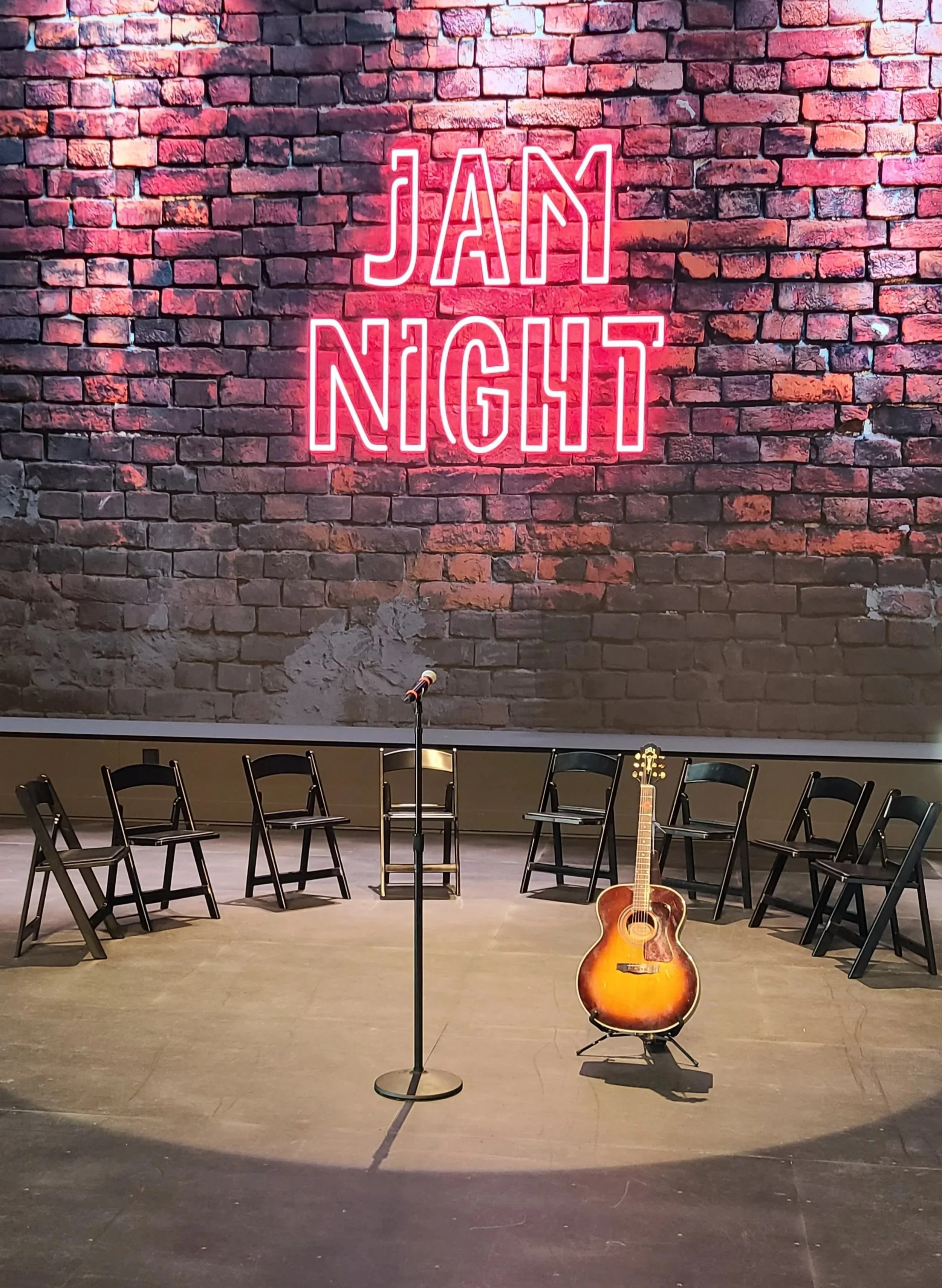 a stage with a semi circle of chairs and a microphone and an acoustic guitar; the wall behind it has a graphic of brick with a neon sign that says "Jam Night"