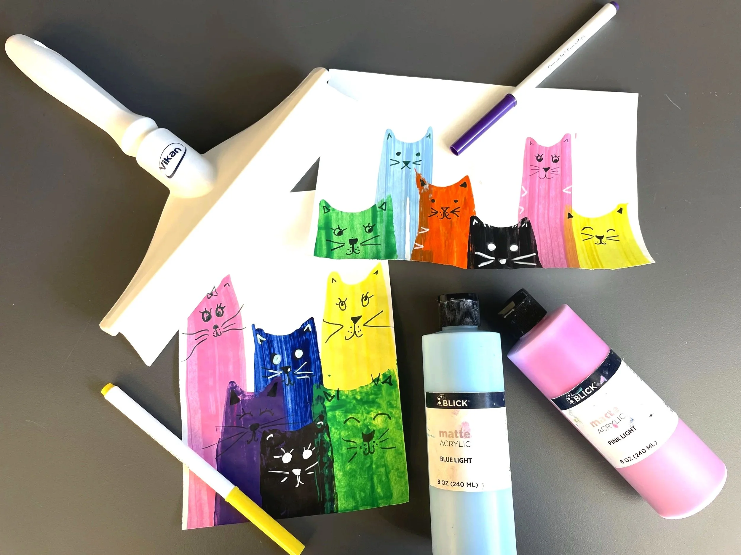 a picture of cat swipe art with colorful painted cats on paper next to paint and pens and a squeegee