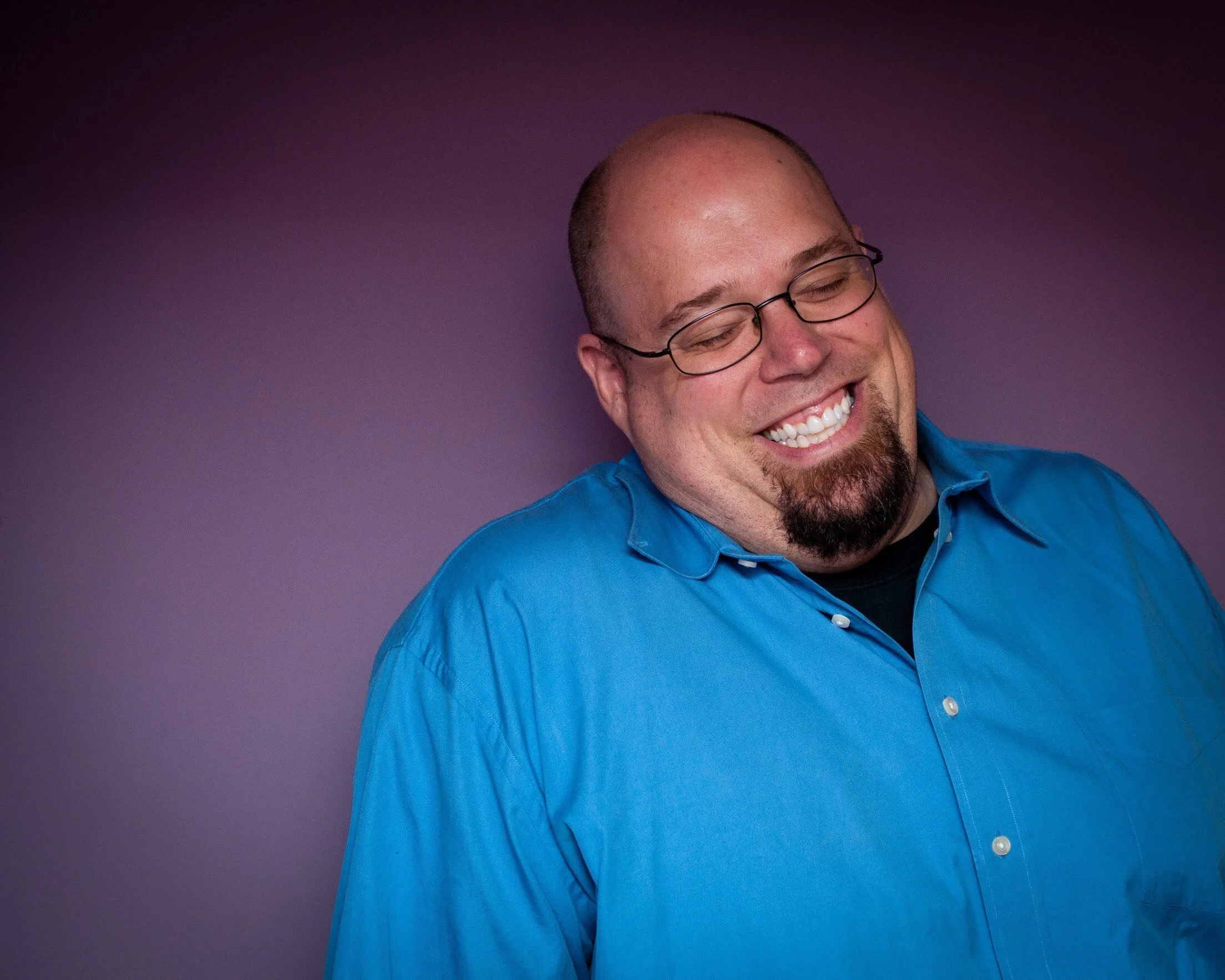 a headshot of comedian Mark Poolos smiling with closed eyes in front of a purple background