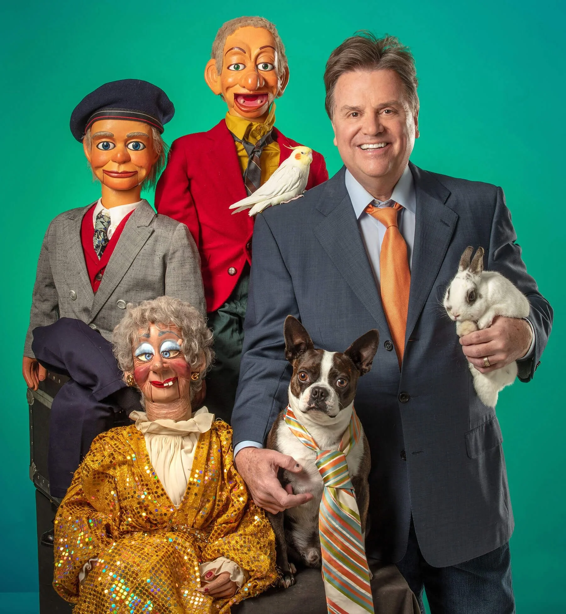Todd Oliver the ventriloquist standing next to several "dummies" and holding a rabbit with a bird on his shoulder and Irving the "talking dog" next to him