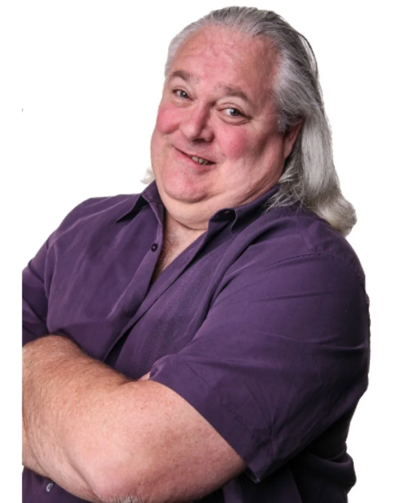a headshot of John Russell with arms crossed wearing a purple short-sleeve shirt and he has long gray hair