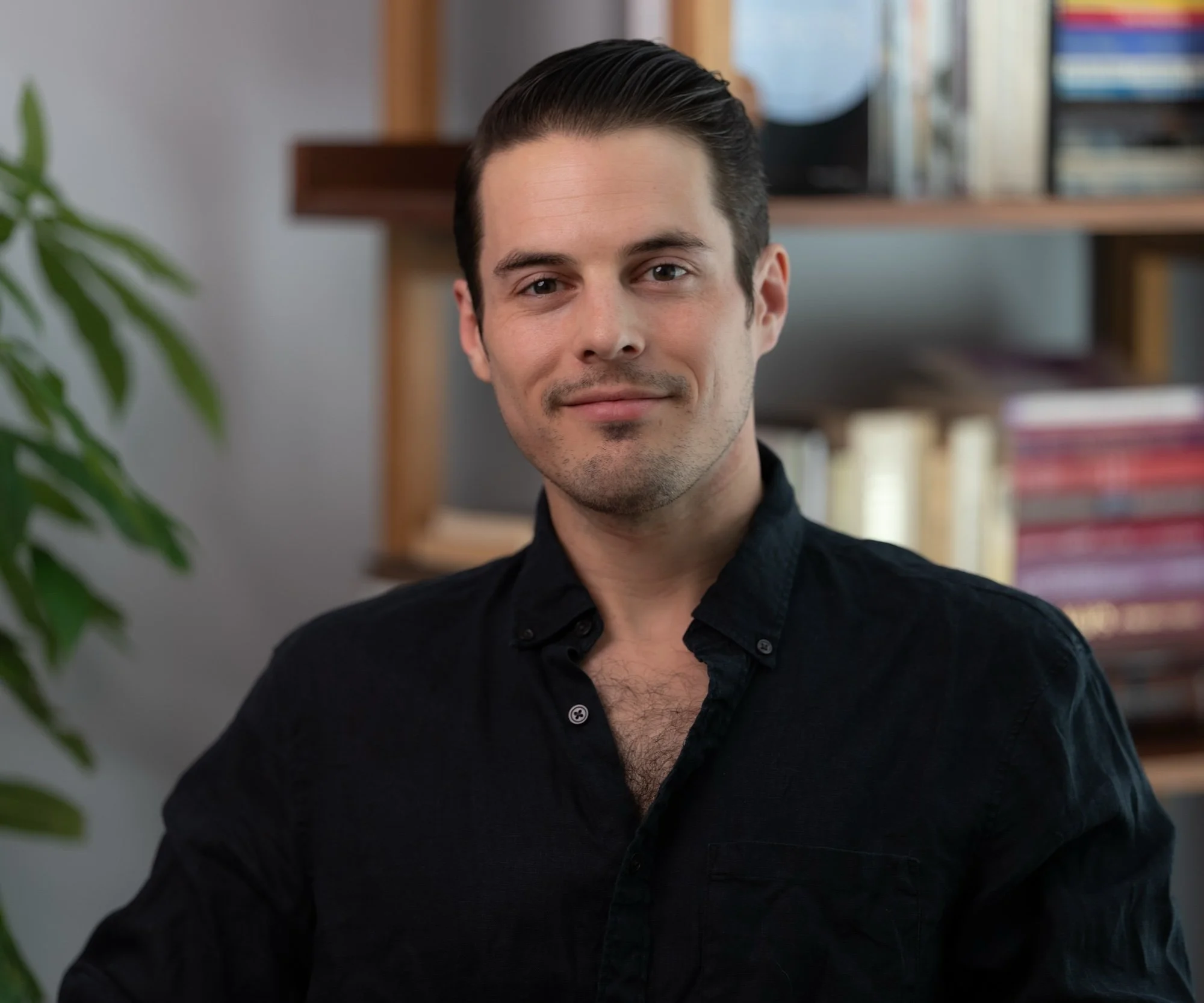 A close-up of a young man with short dark hair, blue eyes, light skin, and a slight smile, wearing a black shirt. He is in a room with shelves of books.