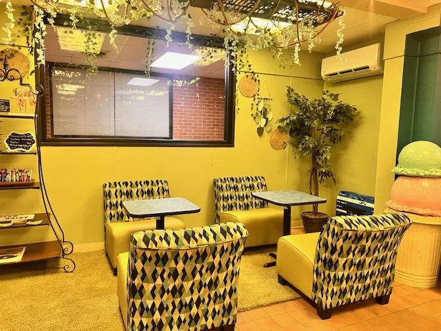 Interior of a colorful cafe with yellow walls and patterned chairs, a window, a shelf with books or decor, and a green tree for decoration.