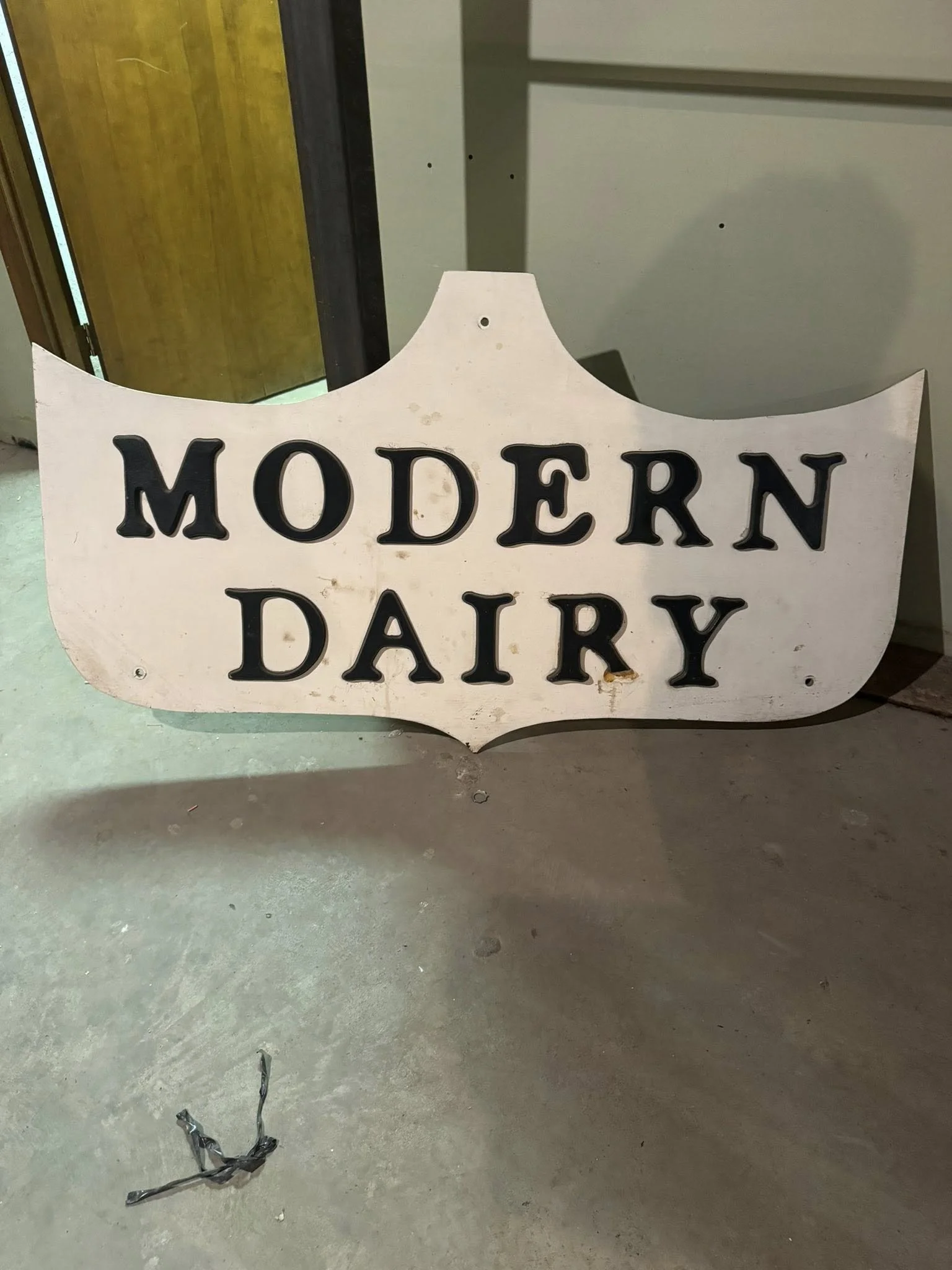 A vintage sign that reads 'Modern Dairy' with black letters on a white background, leaning against a wall on a concrete floor.