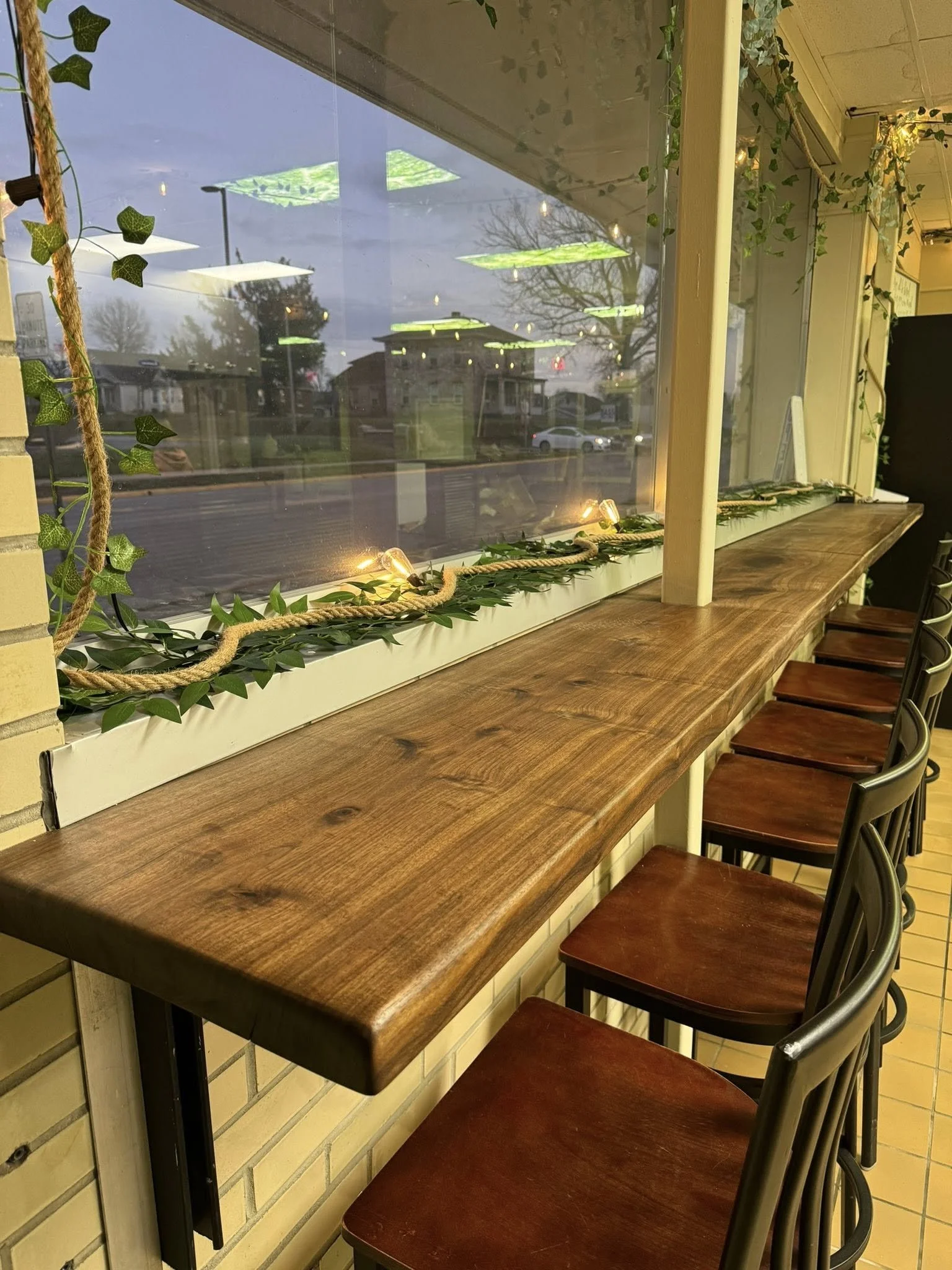 Interior of a café or restaurant with a wooden counter along a large window decorated with green leaves, rope, and string lights; chairs are positioned along the counter.