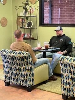 Two men sitting and conversing in a cozy cafe, with drinks on the table and decorative wall art behind them.