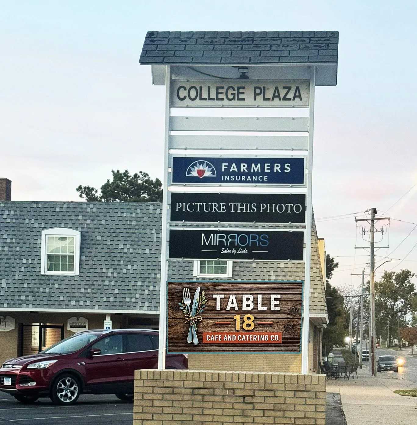 A large vertical sign with multiple business advertisements including College Plaza, Farmers Insurance, a placeholder for a photo, Mirrors Salon by Linda, and Table 18 Cafe and Catering Co., in front of a brick building and parked cars on the street.
