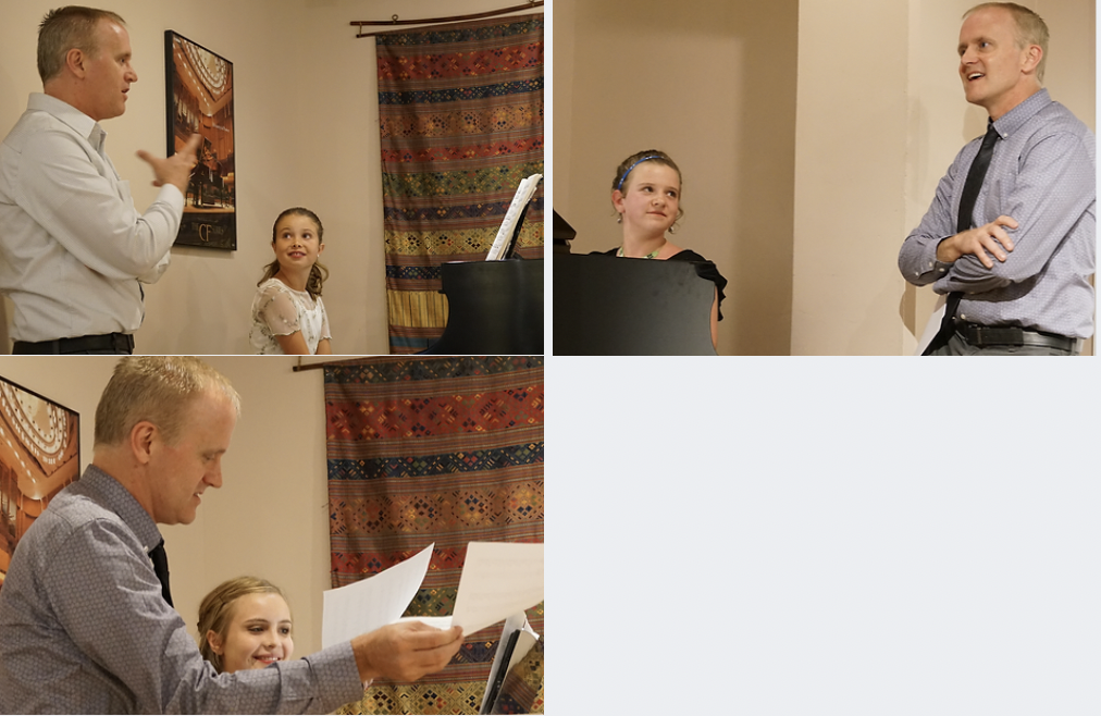 A man talking to a young girl in a room decorated with colorful curtains, with a piano and music sheets nearby, and another man standing and smiling in the background.