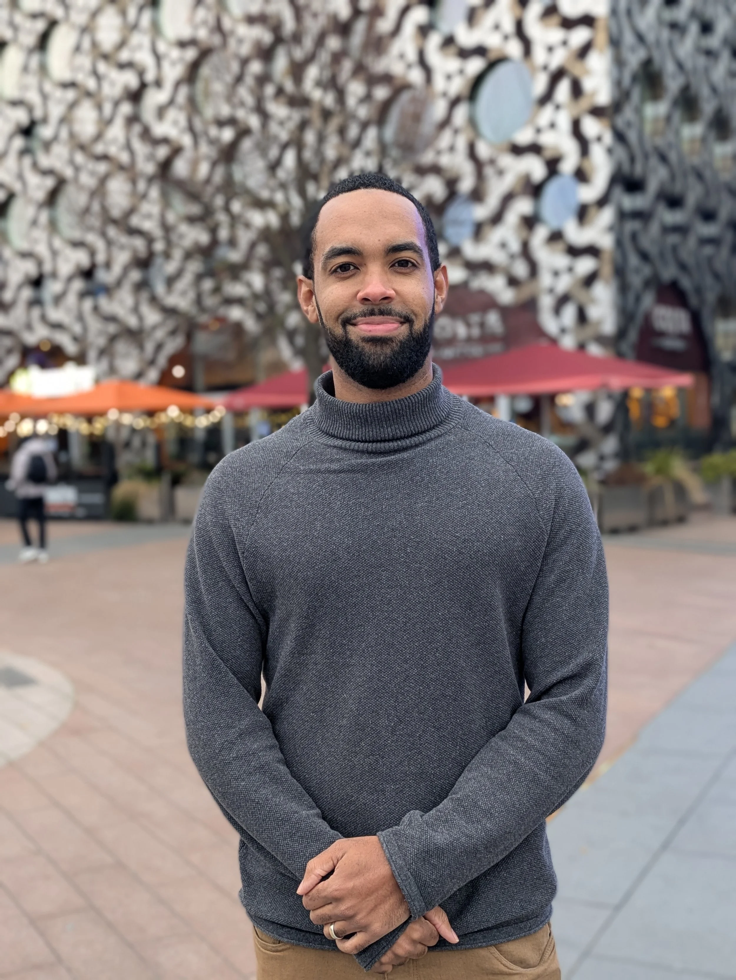 A man with a beard and mustache, wearing a gray turtleneck sweater, standing outdoors in front of a decorative wall with white abstract designs and red umbrellas in the background.