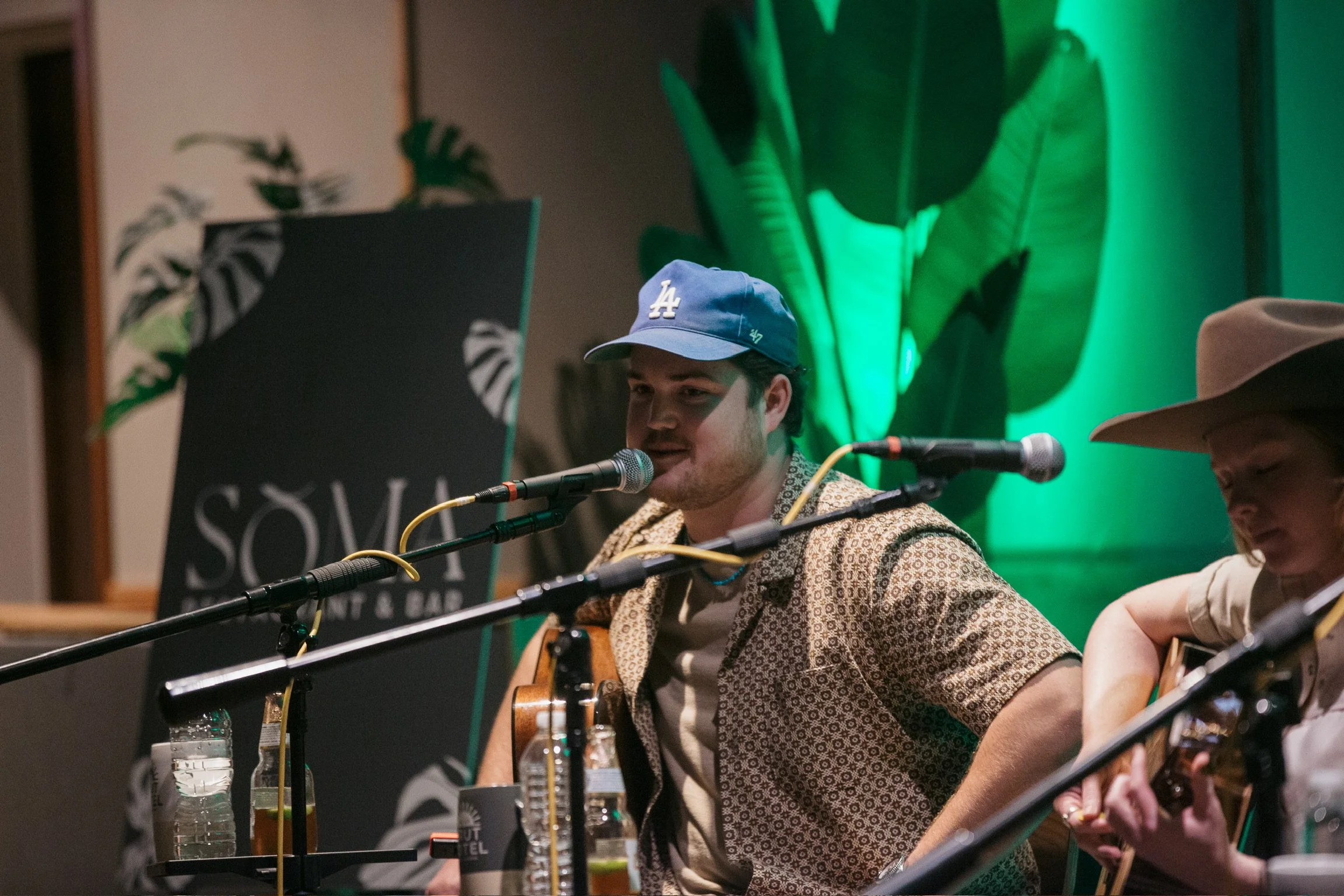 John Ferrell wearing a blue LA baseball cap and patterned shirt singing into a microphone while playing guitar in a green-lit indoor setting, with another person playing guitar beside him.
