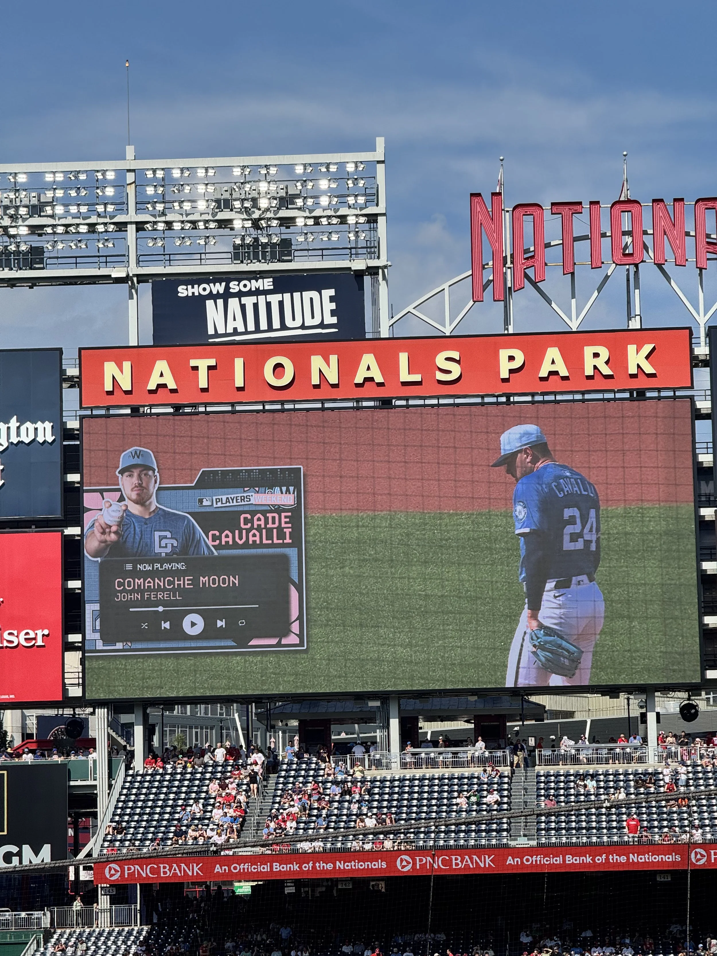 A large digital scoreboard at Nationals Park baseball stadium showing a player in a blue uniform and cap, with a fan base in the background. The screen displays a music player with the song 'Comanche Moon' by John Ferrell playing, and a photo of Cade Cavalli in the corner. The sign above the scoreboard reads 'NATIONALS PARK' in red and yellow.