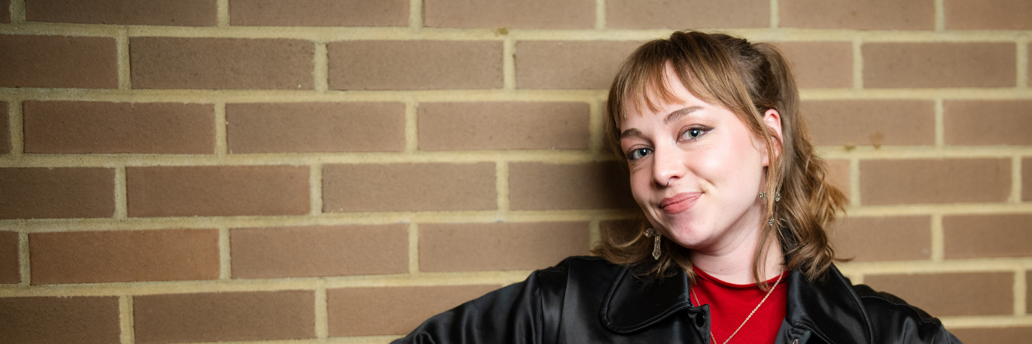 A young woman with shoulder-length blonde hair and green eyes smiling, standing in front of a brick wall.