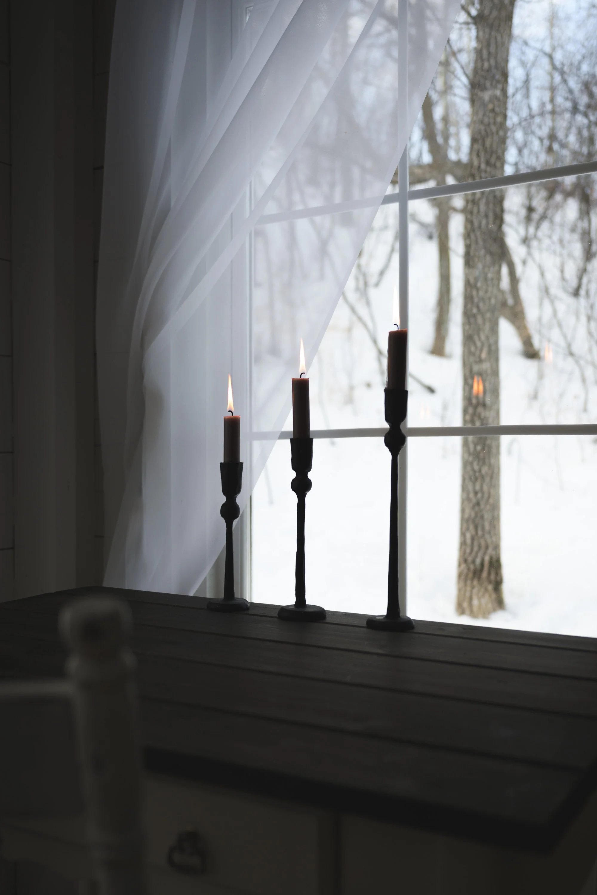 Three lit candles on black candleholders on a wooden surface by a window with sheer white curtains, with snow-covered trees visible outside.
