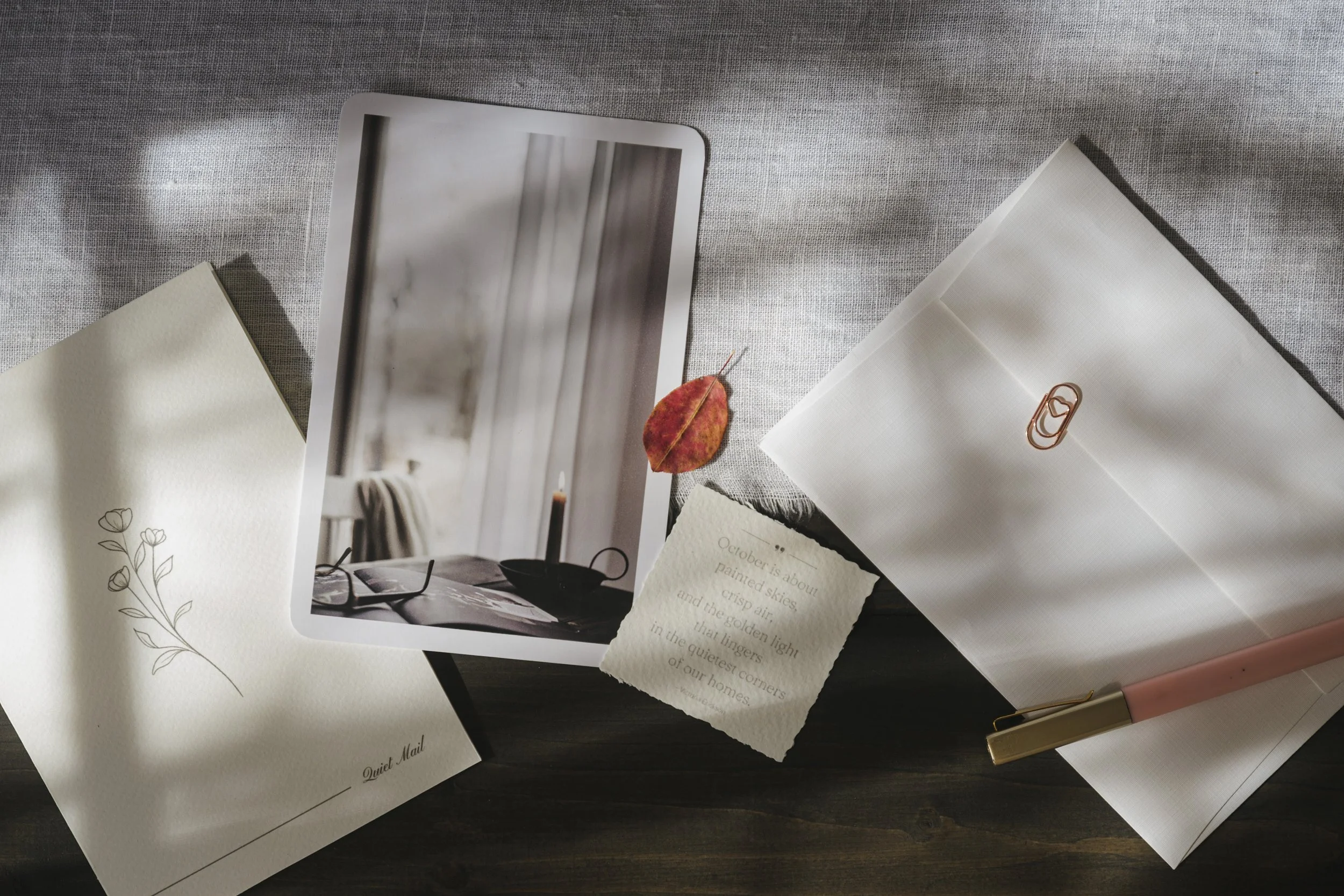 Flat lay of a collection of papers and a photo on a dark surface. The photo depicts a cozy window scene with a cup, a newspaper, and a candle. One paper features a delicate line drawing of flowers, labeled 'Quiet Mail.' Another paper has a poem about October's skies, and a small dry autumn leaf is placed nearby. There is a white envelope with a copper paperclip, a pink pen with gold accents, and some fabric underneath.