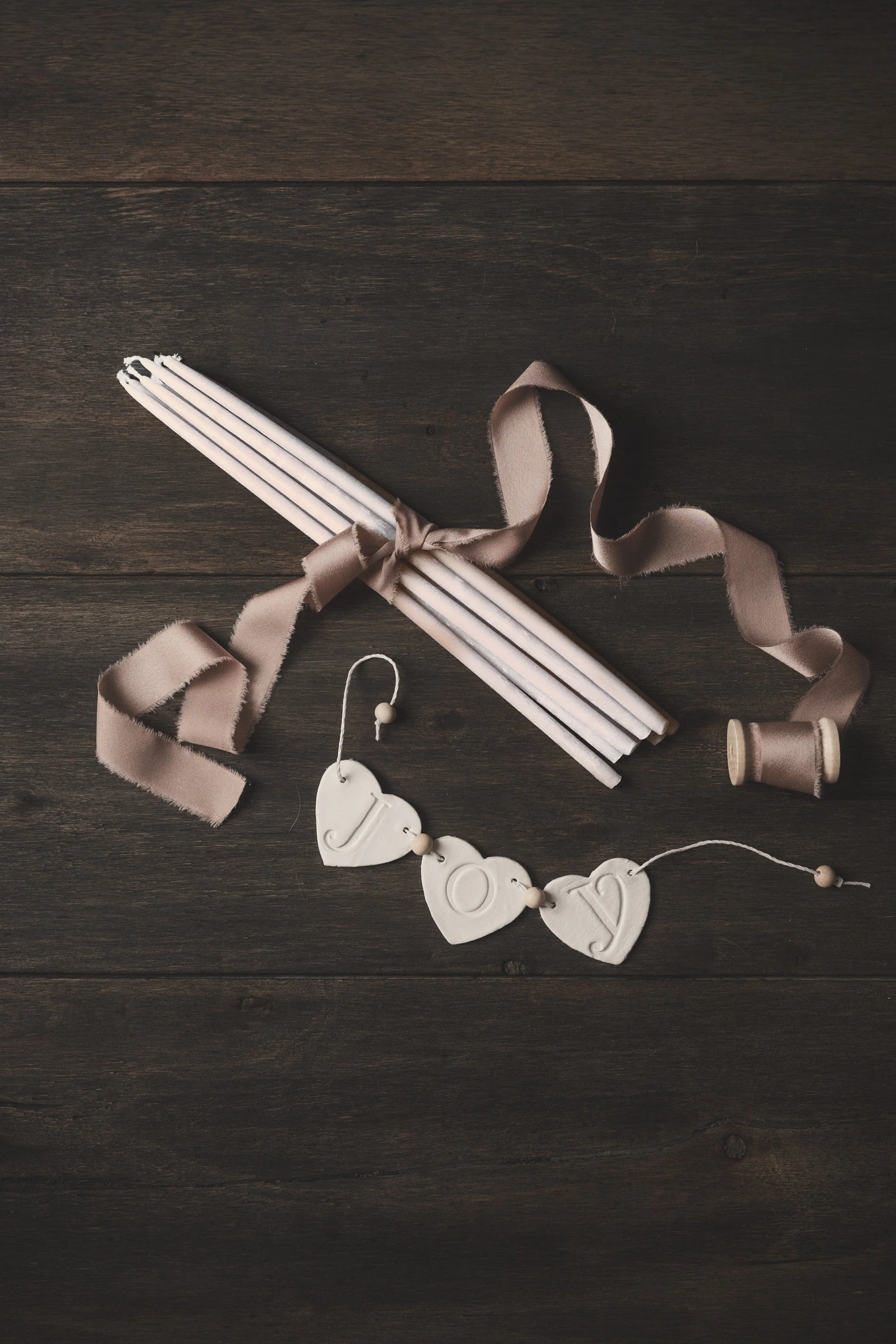 White candles tied with mauve ribbon beside heart-shaped joy garland on dark wood