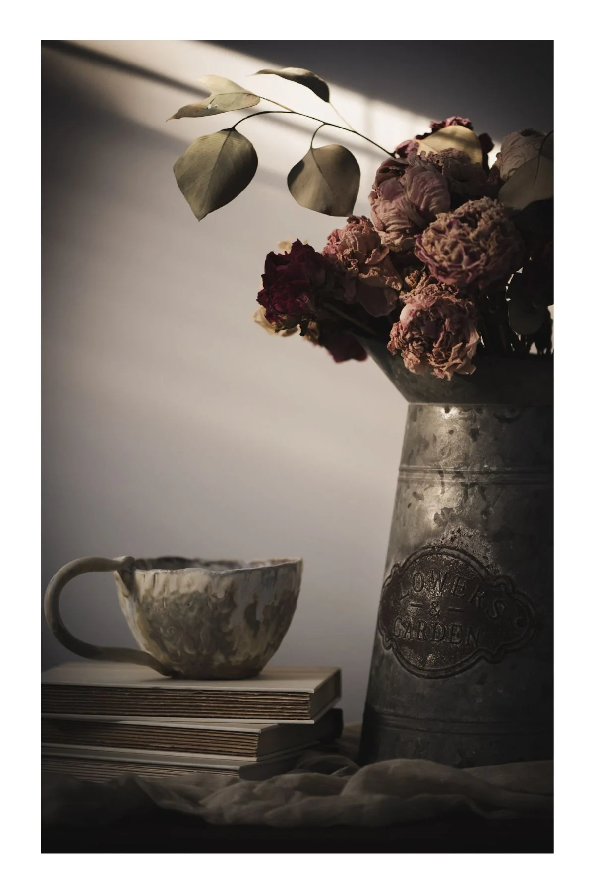 A still life scene with a bouquet of dried pink and red flowers in a gray metal milk can, a ceramic cup with a floral pattern, and a stack of two books on a dark surface, illuminated by soft natural light.