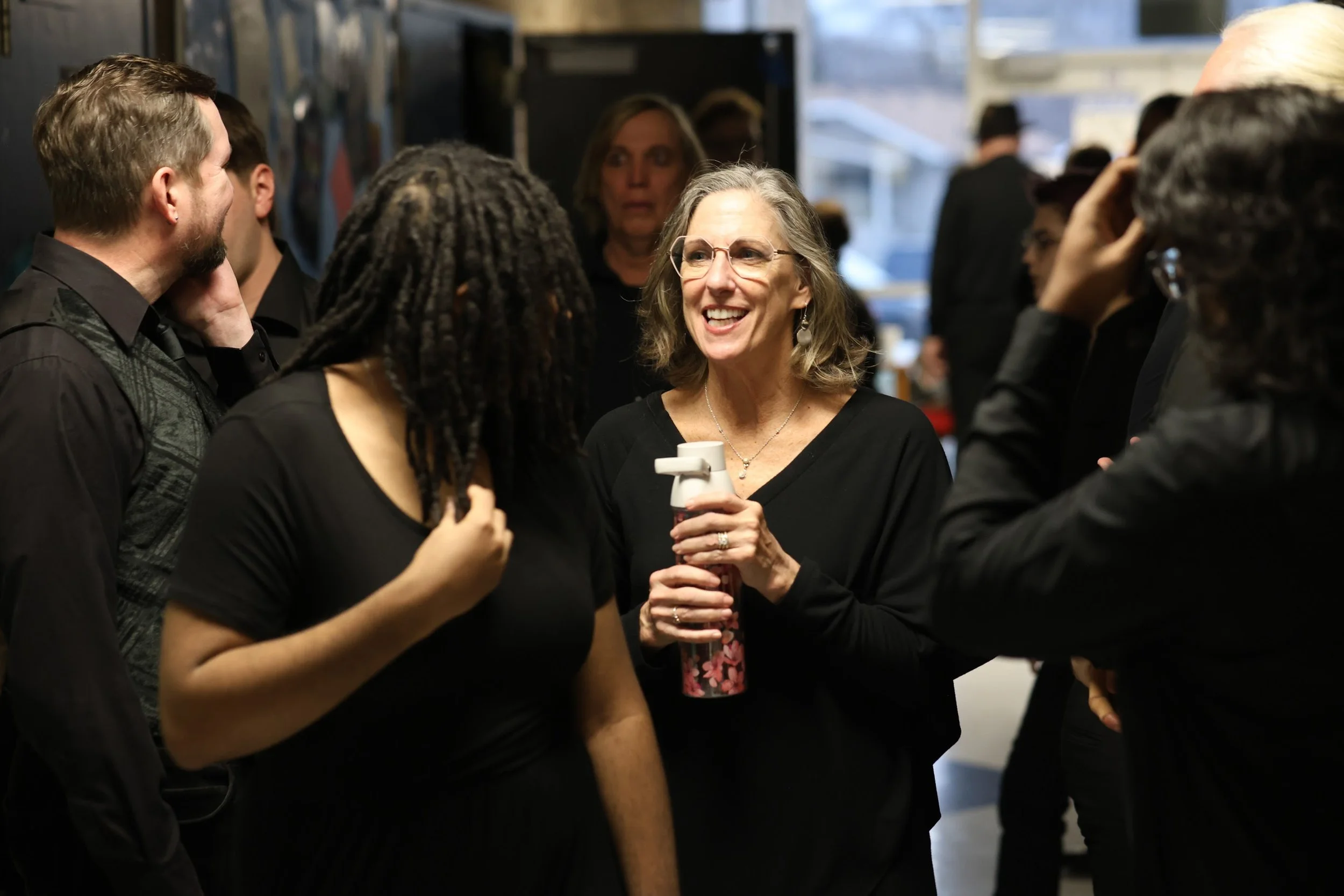 A group of people conversing in a social setting, with a smiling elderly woman holding a spray bottle as a prop.