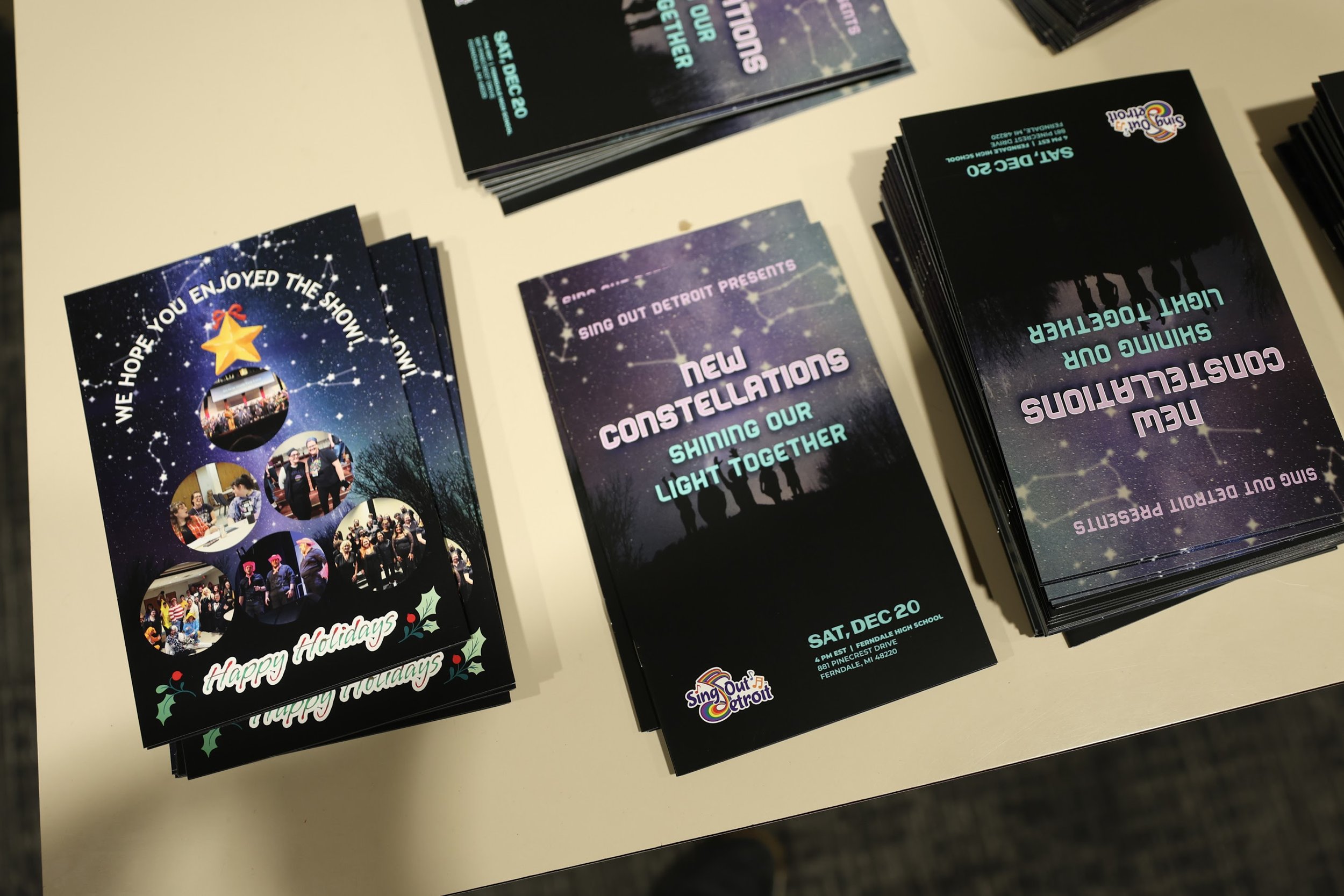 Brochures and flyers on a table advertising a holiday event called 'New Constellations.' One flyer features holiday-themed photos, a Christmas tree ornament, and the message 'Happy Holidays.' The other flyers promote an event titled 'Shining Our Light Together' scheduled for Saturday, December 20, at Ferndale High School, presented by Sing Out Detroit.