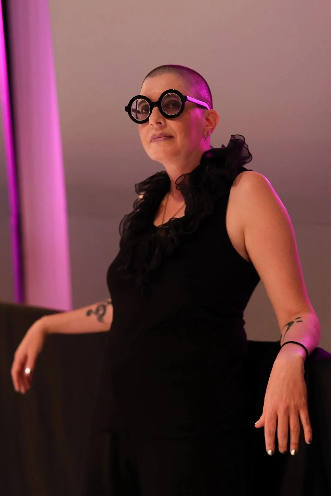 A woman with a shaved head wearing black sunglasses with circular frames, a sleeveless black top with ruffled details around the neckline, and tattoos on her arms, standing against a neutral background with purple lighting.