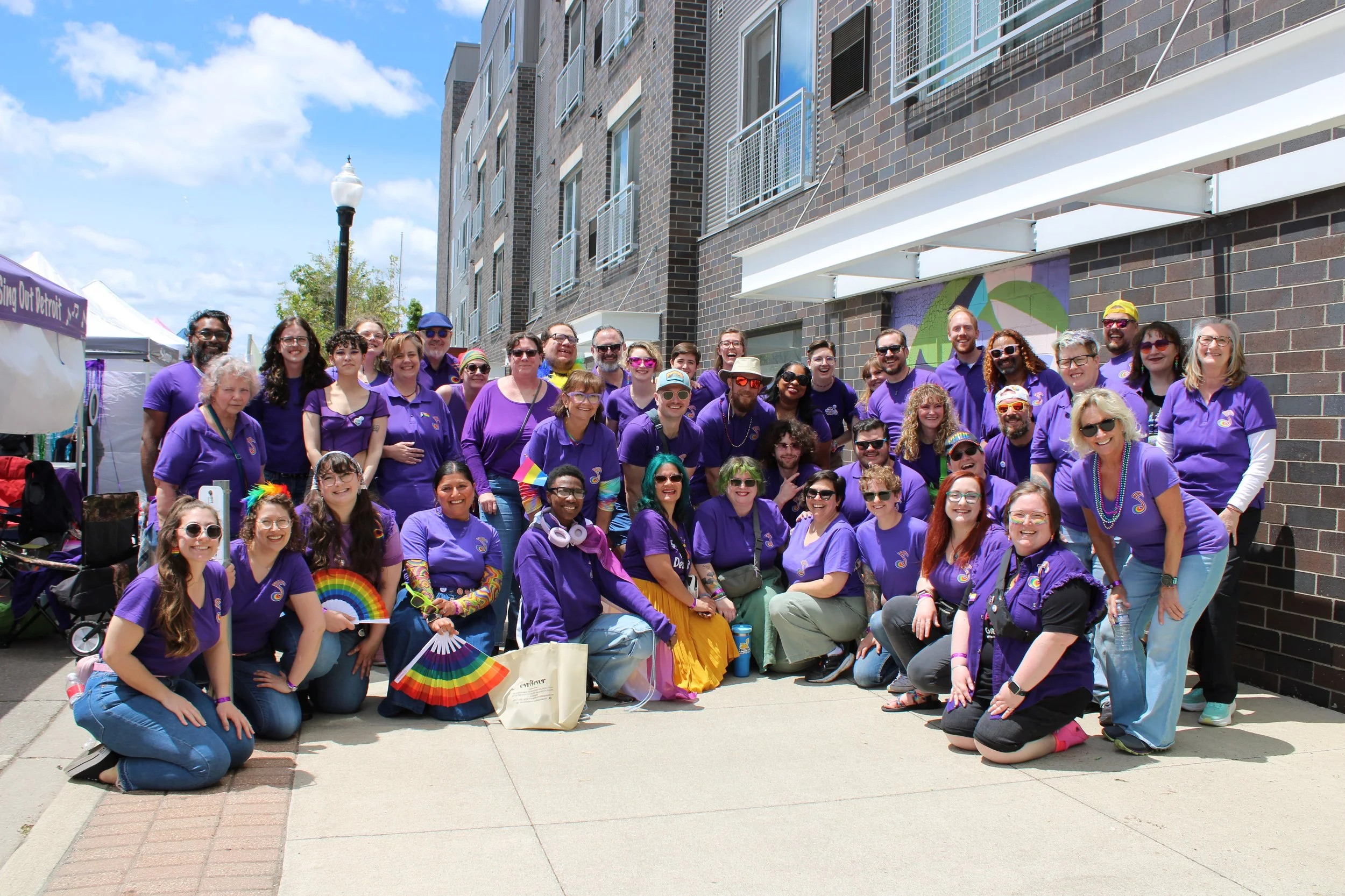 Group of diverse people gathered outdoors, many wearing purple shirts and accessories, celebrating at a pride event on a sunny day with blue sky and clouds, in front of a brick building.