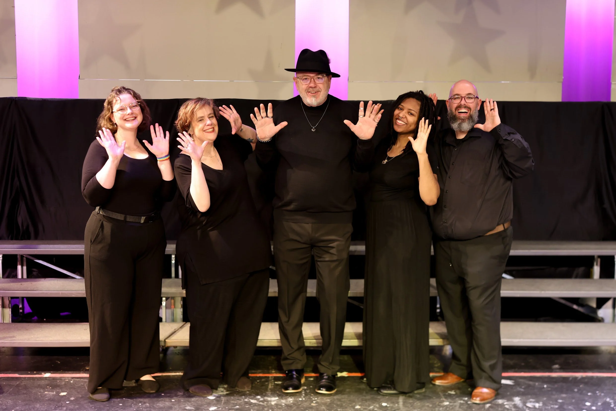 Group of five smiling people standing on a stage, raising their hands, dressed in black, with a backdrop of purple and pink columns and stars.