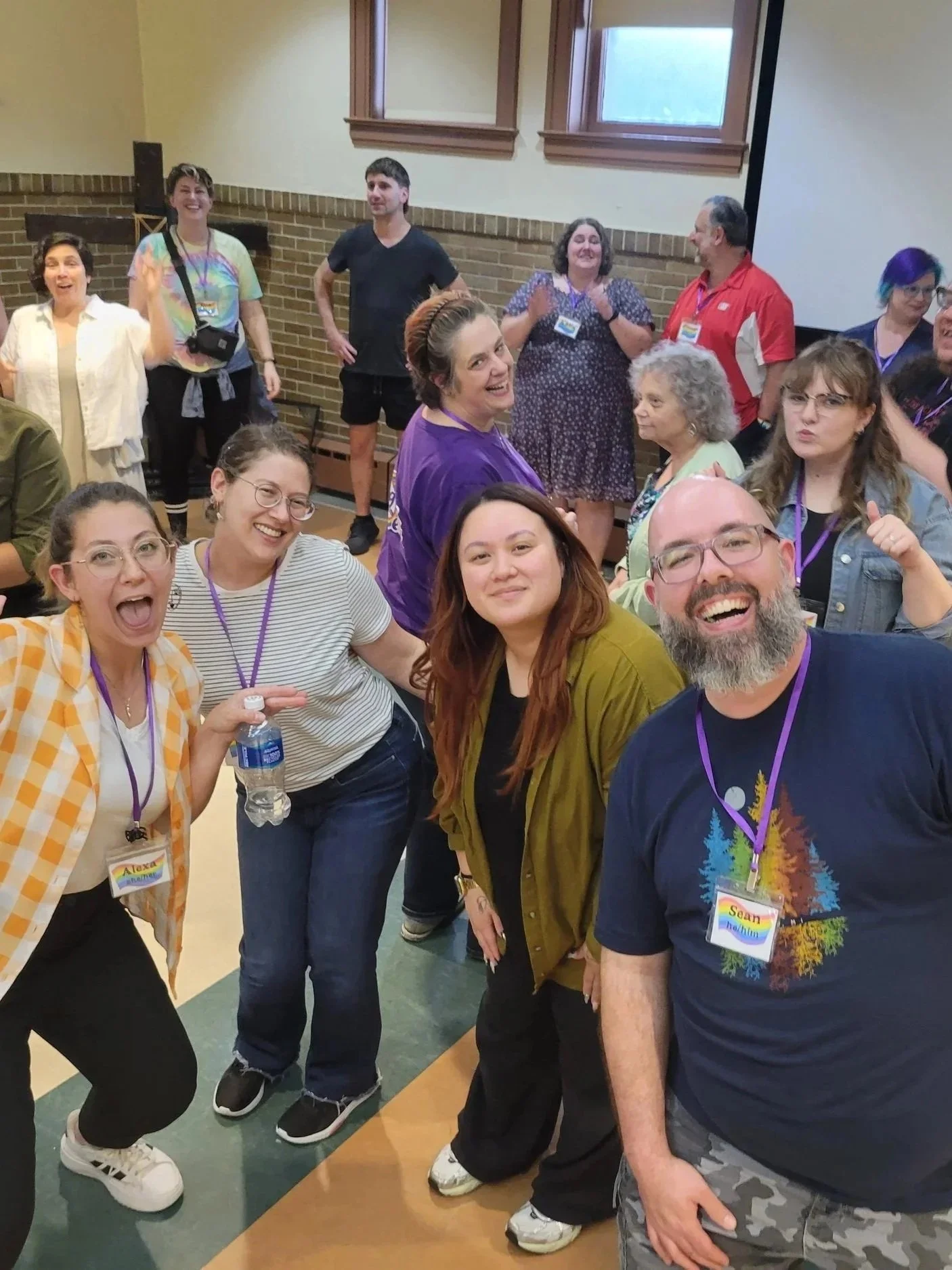 Group of smiling people gathered in a room, some wearing name tags, with a brick wall and windows in the background.