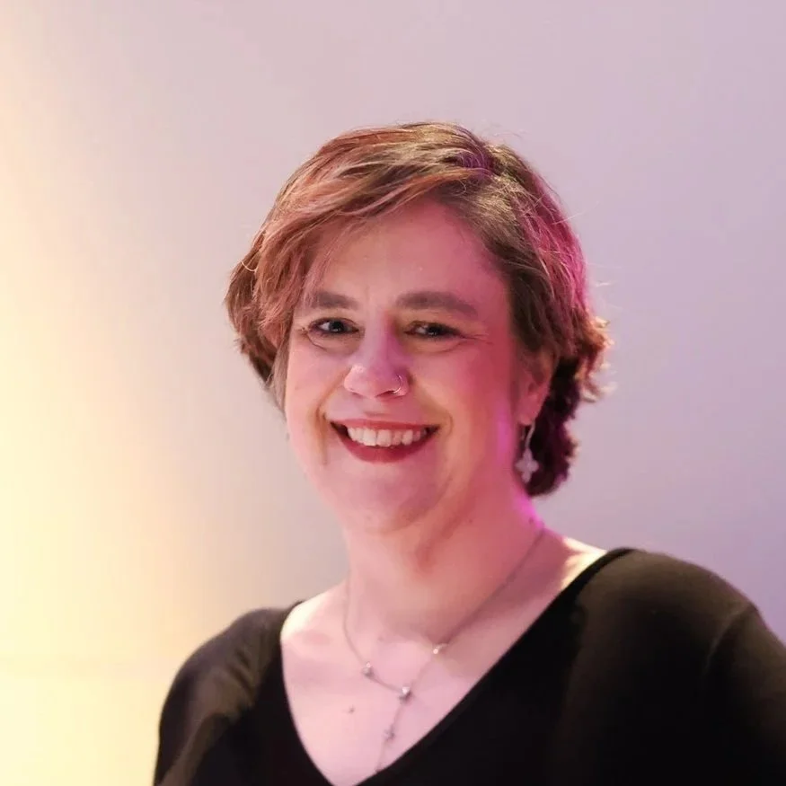 A person with short, light brown hair smiling and winking, wearing a black top, earrings, a nose ring, and layered necklaces against a plain background.