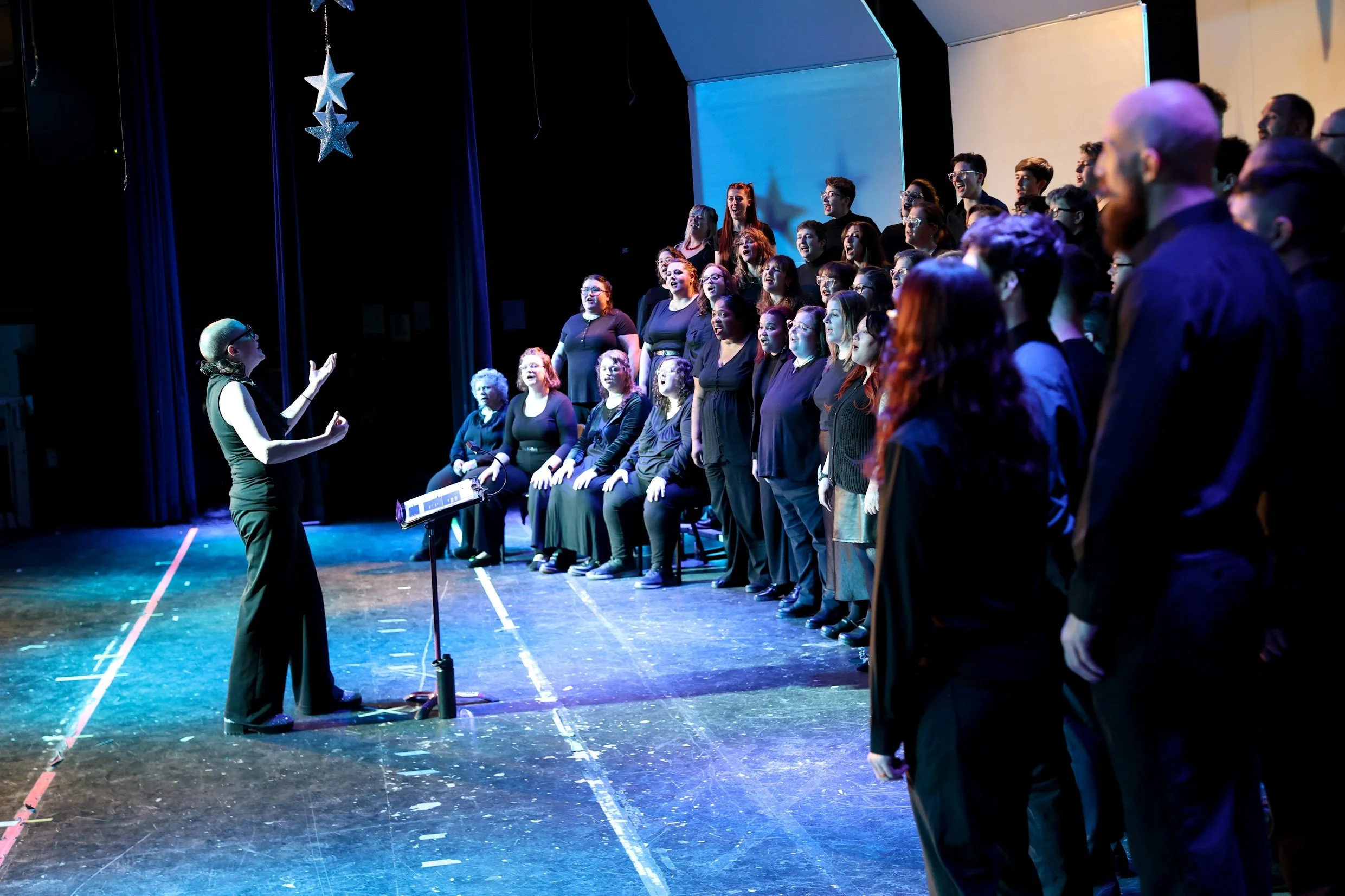 A choir performing on stage with a conductor in front, and stage decorations including hanging stars.