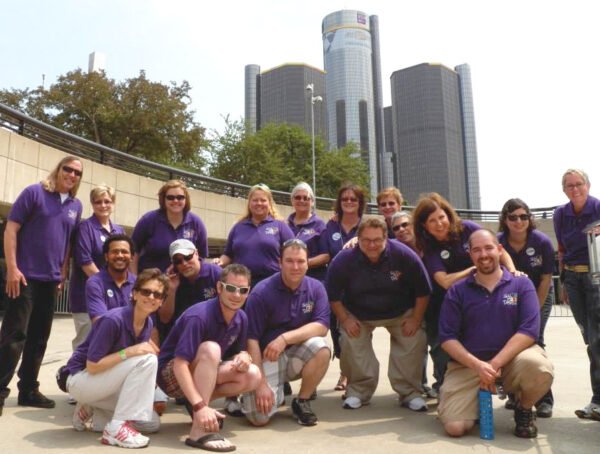 Group of people posing outdoors in front of a cityscape with tall buildings, some wearing purple shirts.