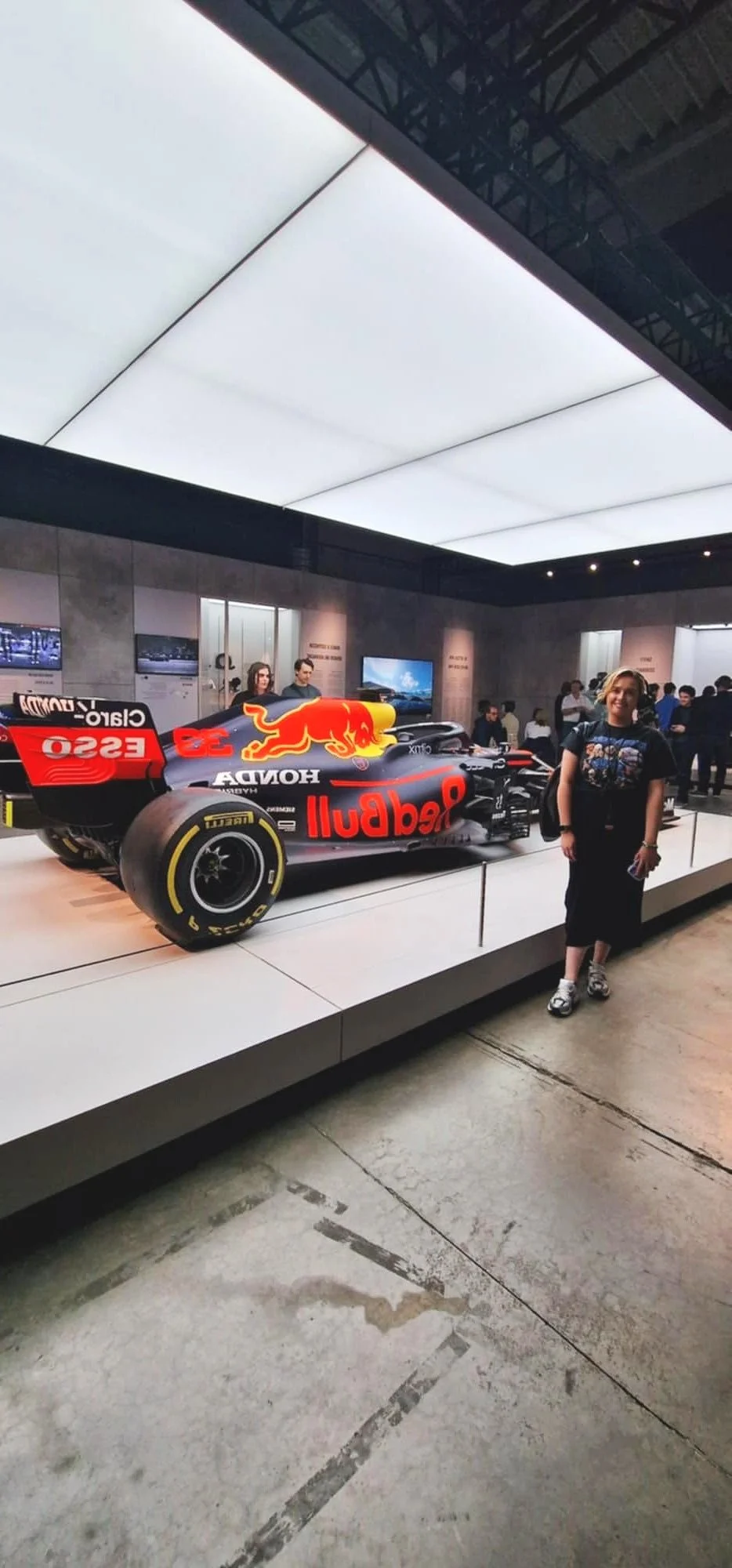 A person standing next to a Red Bull Racing Honda Formula 1 car on display in a showroom or museum, with several other visitors and screens in the background.