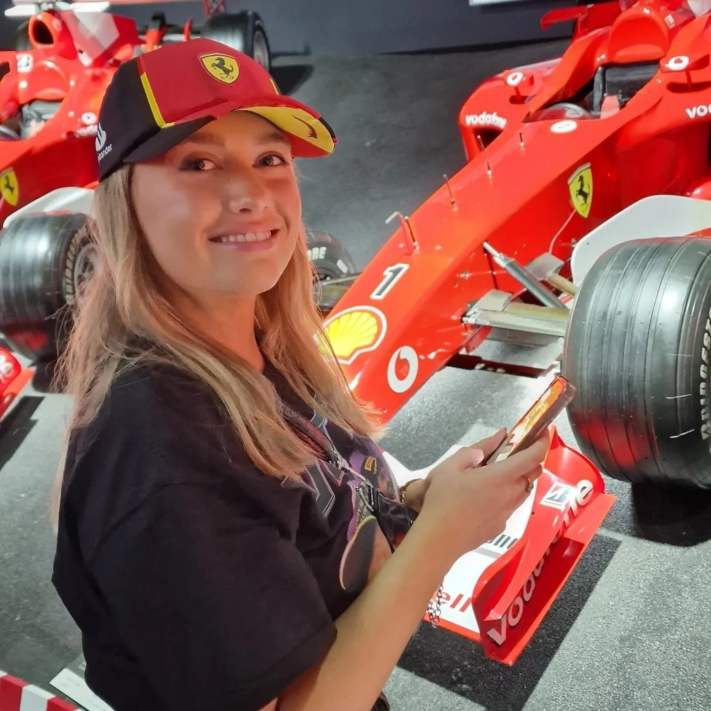 A woman in a Ferrari cap smiling and looking at her phone, standing next to a red Formula 1 race car with Ferrari and Vodafone branding.