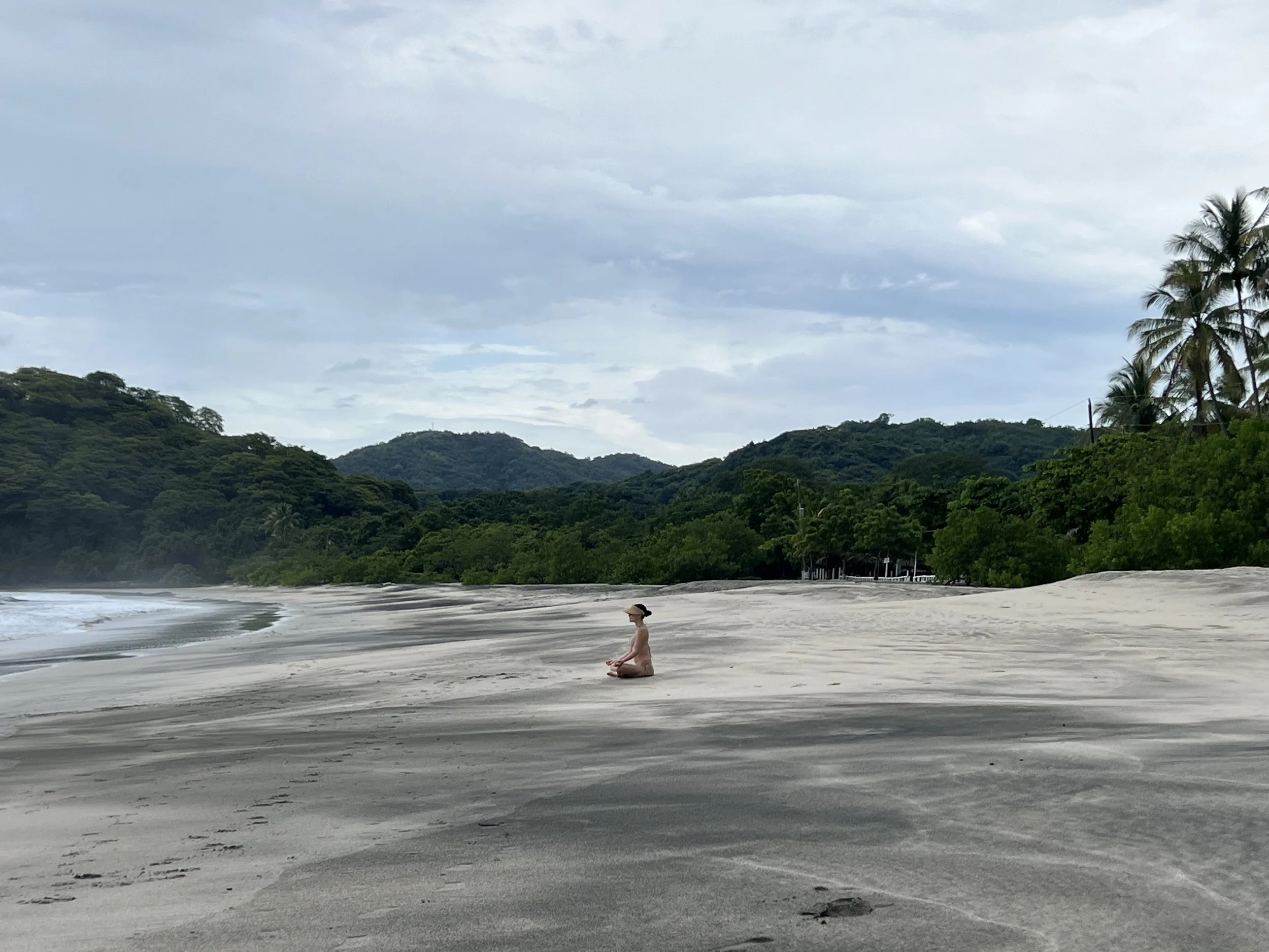 Person sitting on a secluded, sandy beach with lush green hills in the background. The sky is partly cloudy, and palm trees border the beach.