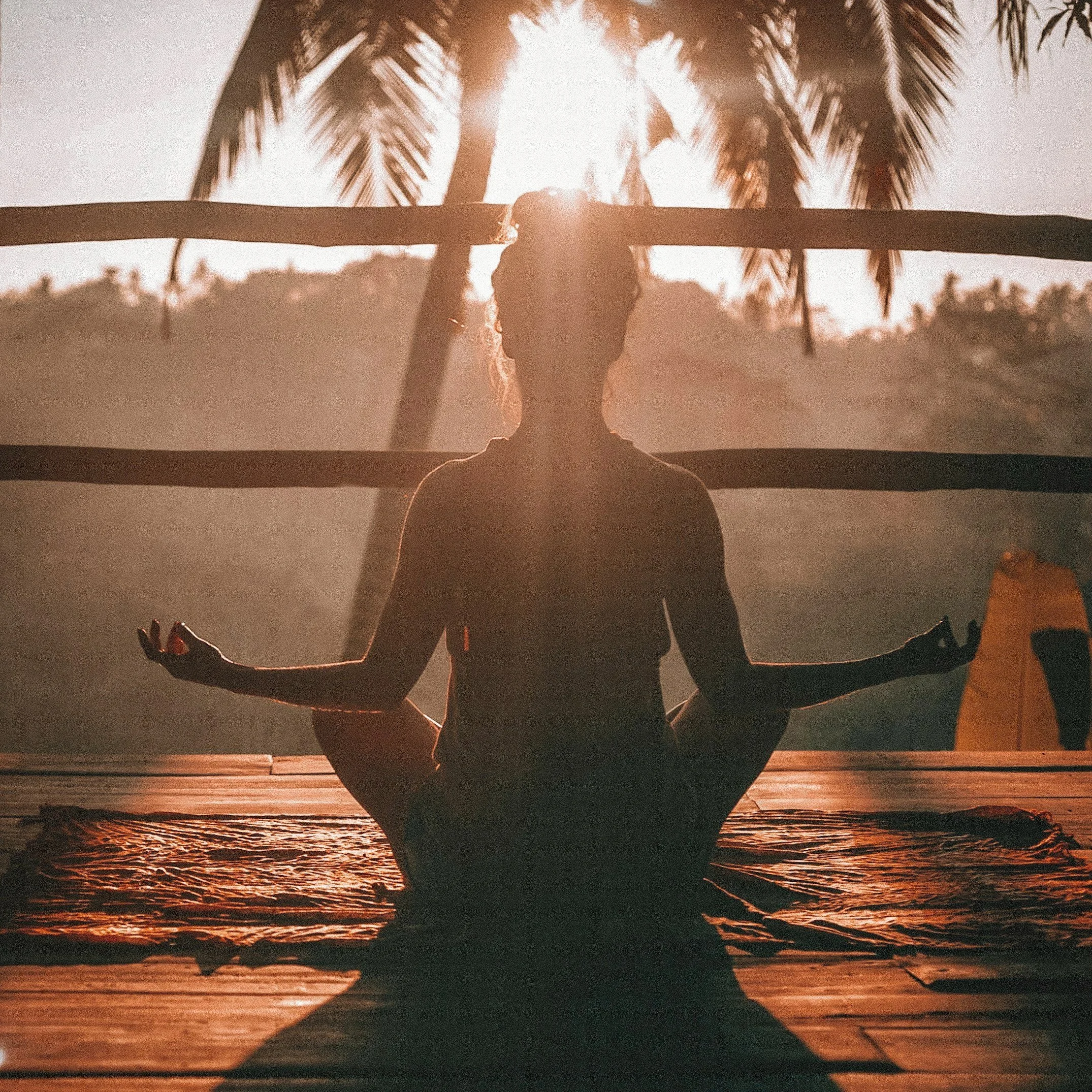 Silhouette of a person meditating in lotus position on a wooden deck at sunrise, with palm trees in the background.
