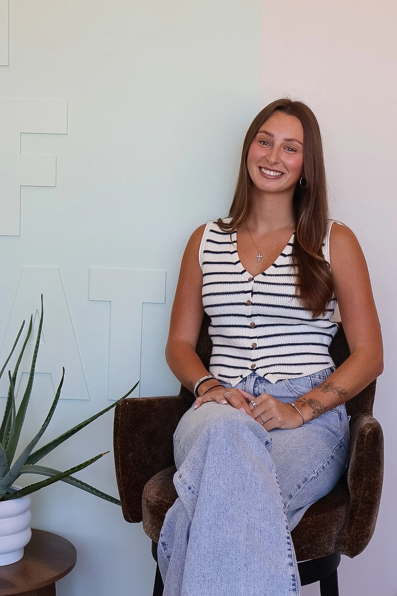 A young woman with long brown hair, wearing a striped sleeveless top and light blue jeans, is sitting in a brown armchair. She is smiling, and wearing jewelry including a cross necklace, hoop earrings, and rings. A potted plant is partially visible on the left. The background is a plain white wall with faint lettering.