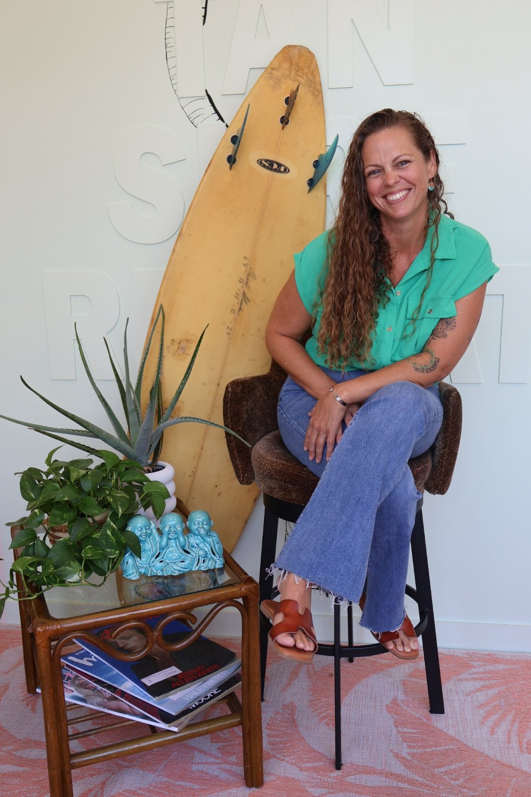 A woman with long curly hair sitting on a chair, smiling, wearing a turquoise shirt and jeans. Behind her is a yellow surfboard and a wall with large white outlined words. To her left are plants, a small table with magazines, and decorative figurines.