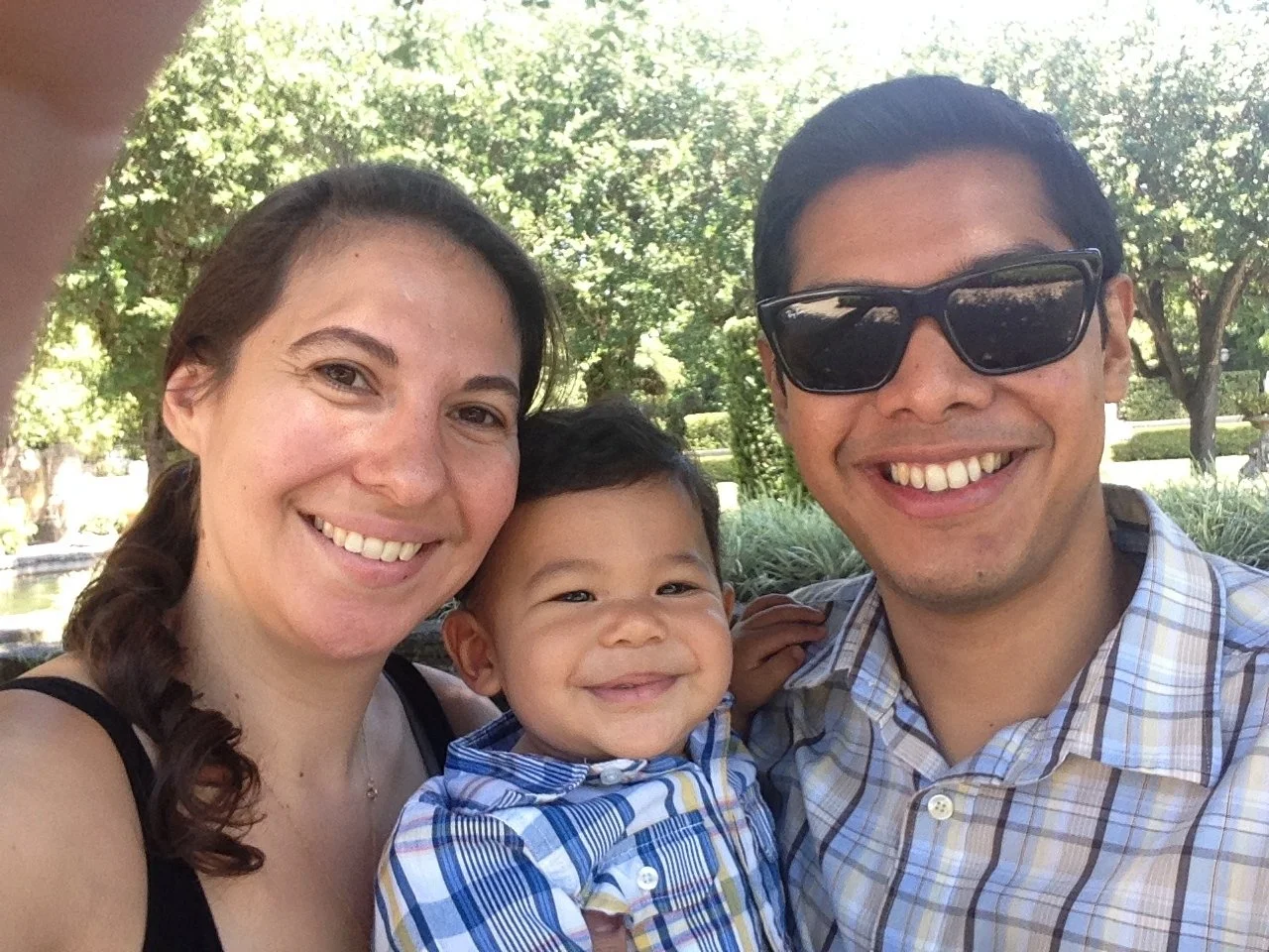 A family of three smiling outdoors with trees in the background, a woman, a man wearing sunglasses, and a young boy.